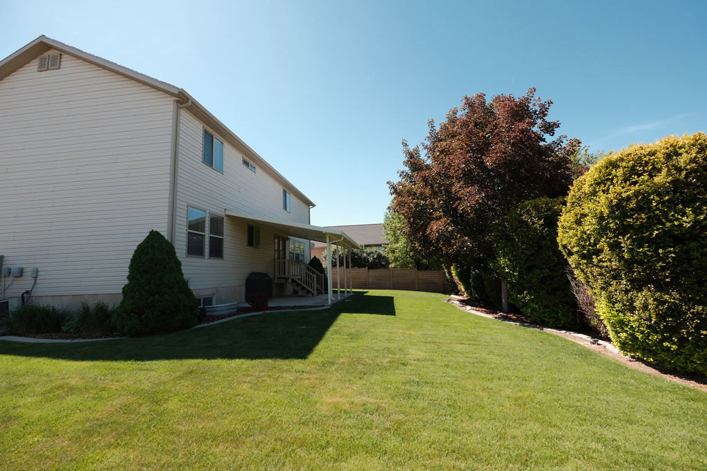Expansive backyard featuring a large green lawn, mature trees and shrubs along the perimeter, and a covered patio with direct access to the residence