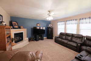 Carpeted living area featuring a corner fireplace with a light-toned surround and hearth, multiple windows with blinds, a ceiling fan, and a painted accent wall