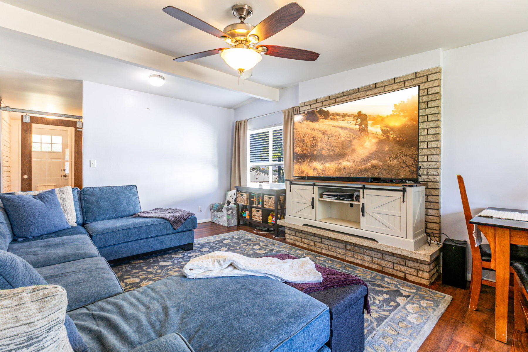 Family area featuring a brick fireplace, wood-like flooring,  and a ceiling fan with integrated lighting