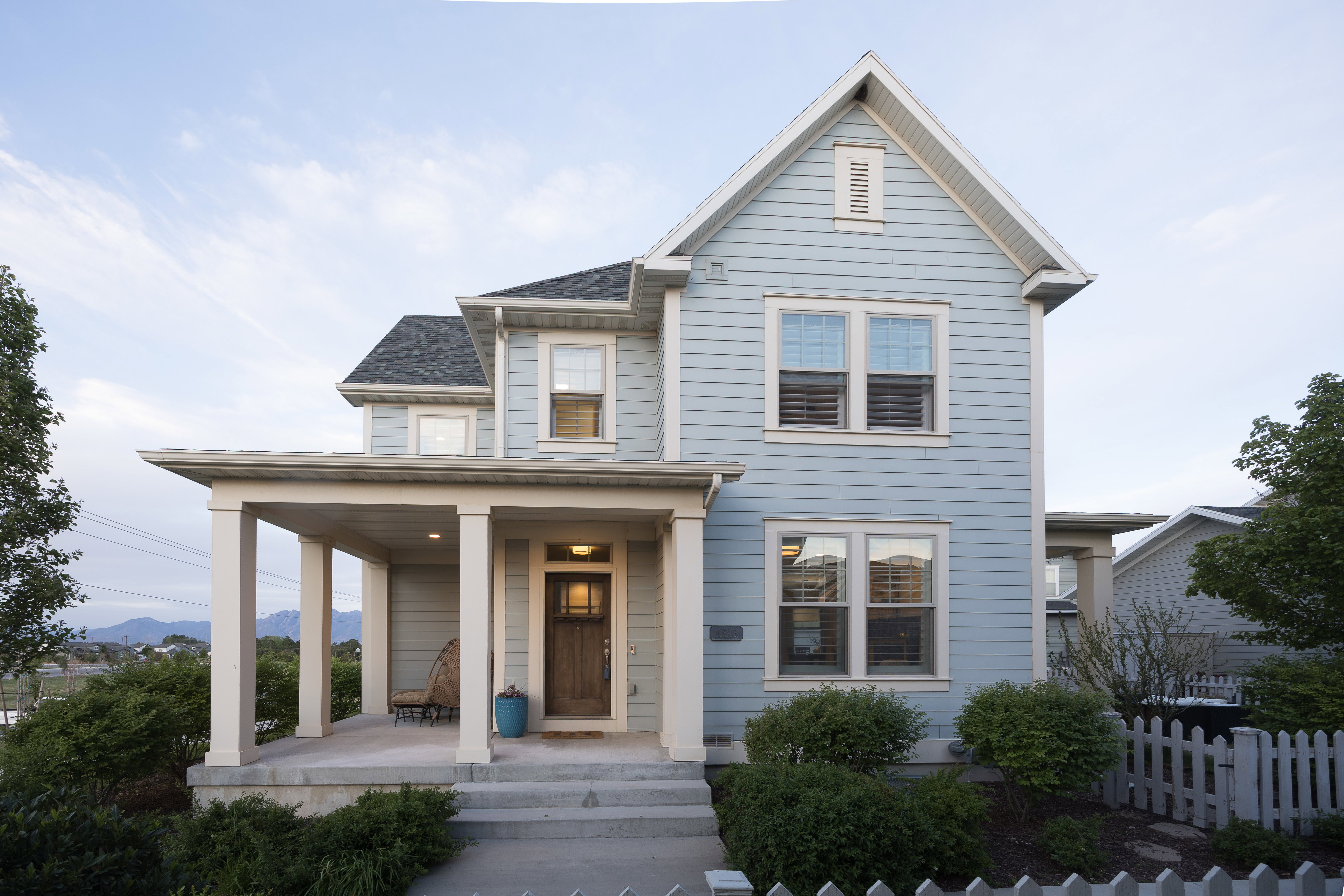 Light blue siding exterior featuring a covered front porch with substantial columns, a dark wood entry door, a gabled roofline, and a white picket fence