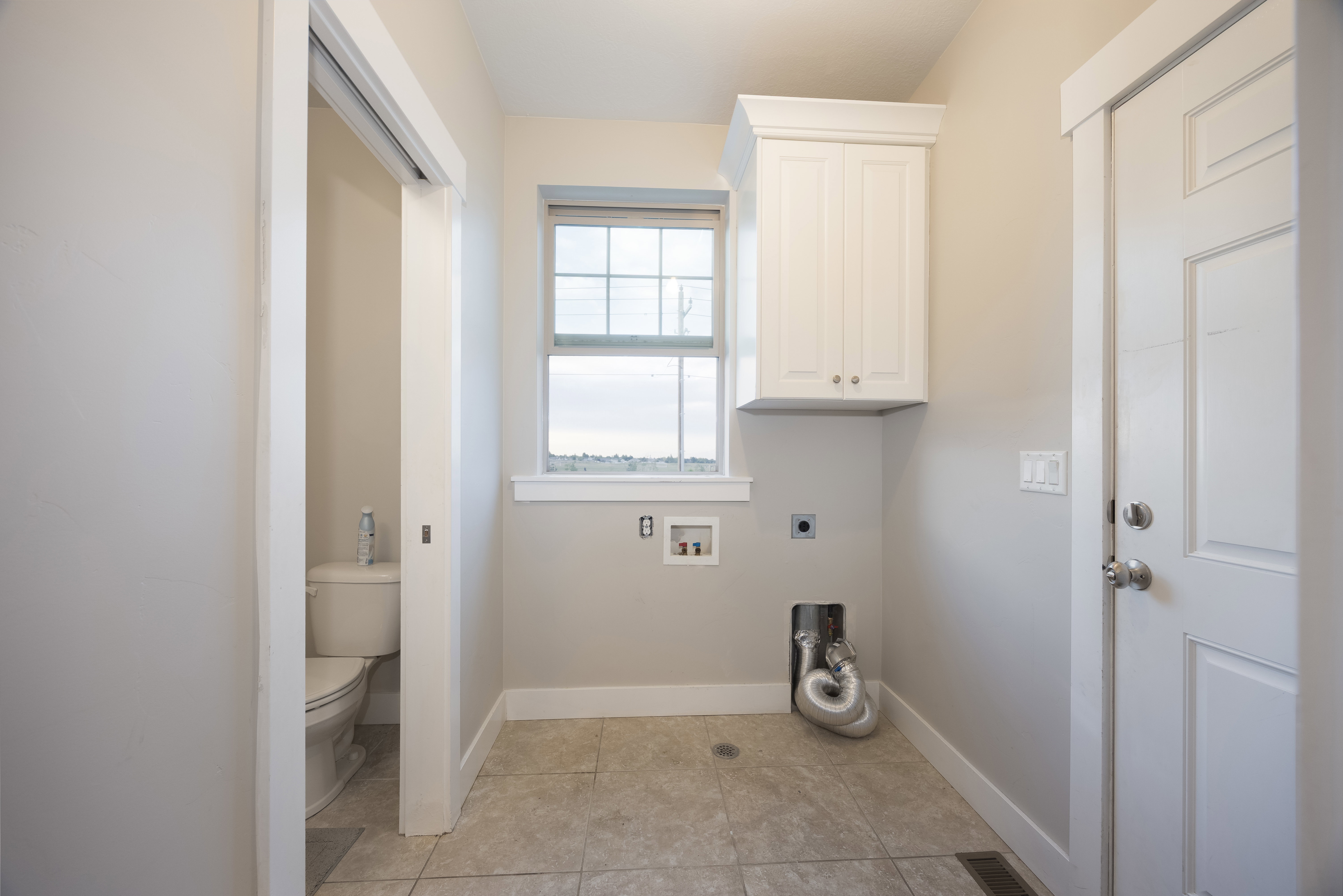 Utility room featuring a window with grid pattern, a white shaker-style wall cabinet, light-toned wall paint, white trim, and light-toned floor tiles