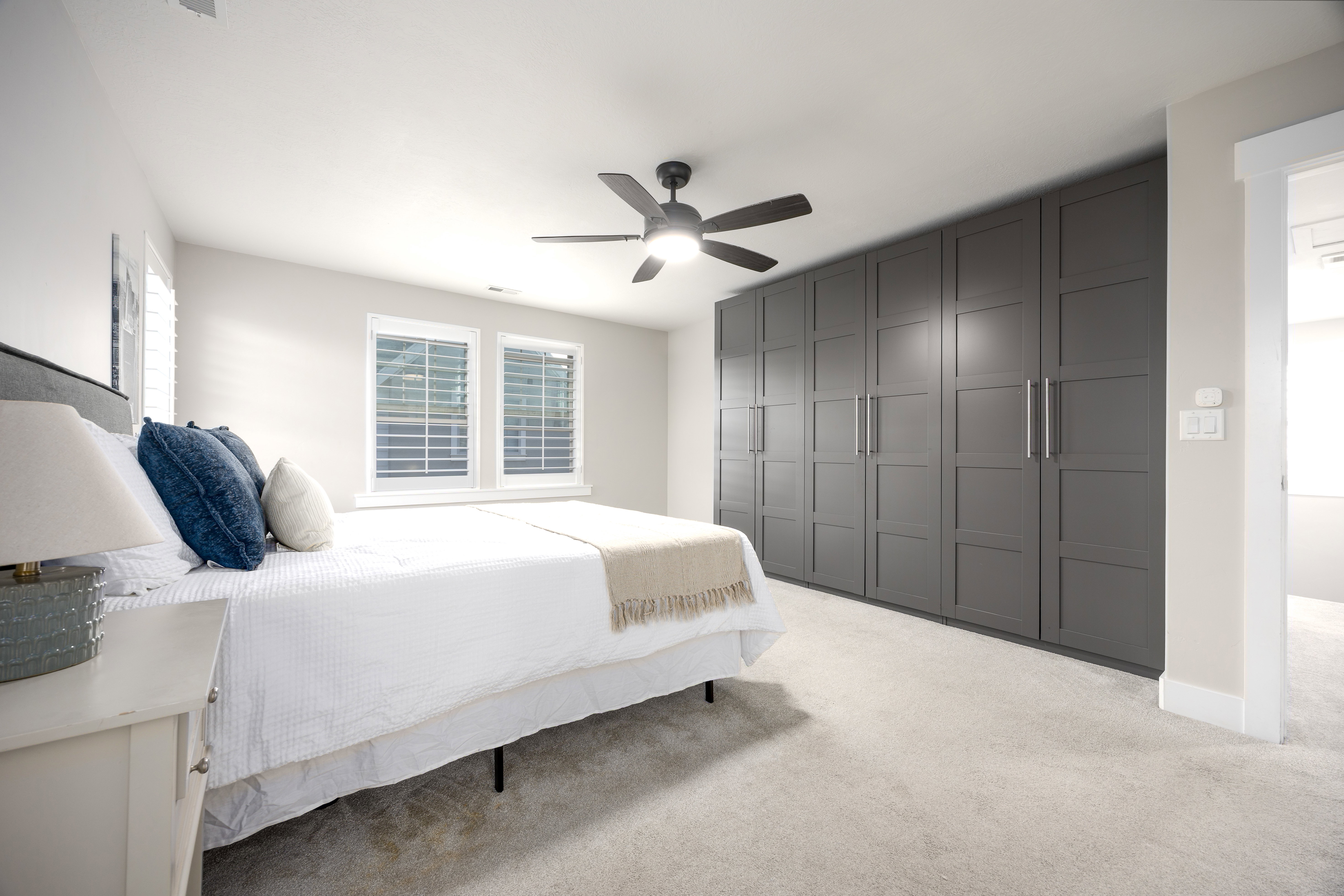 Bedroom featuring extensive built-in gray cabinetry, light-colored carpeting, two windows with white plantation shutters, and a ceiling fan with integrated lighting