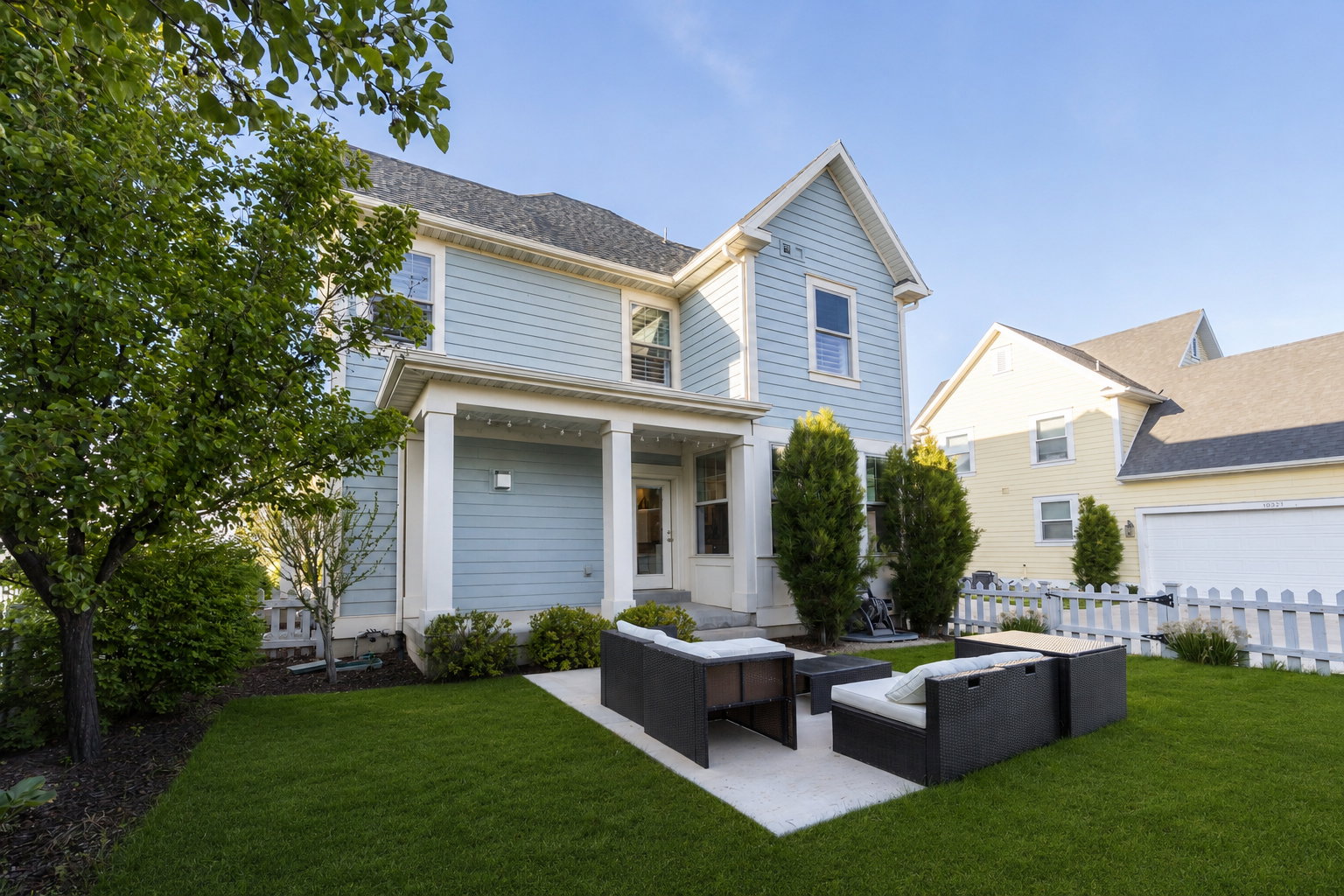 Expansive backyard featuring a concrete patio, manicured lawn, established landscaping, and a white picket fence