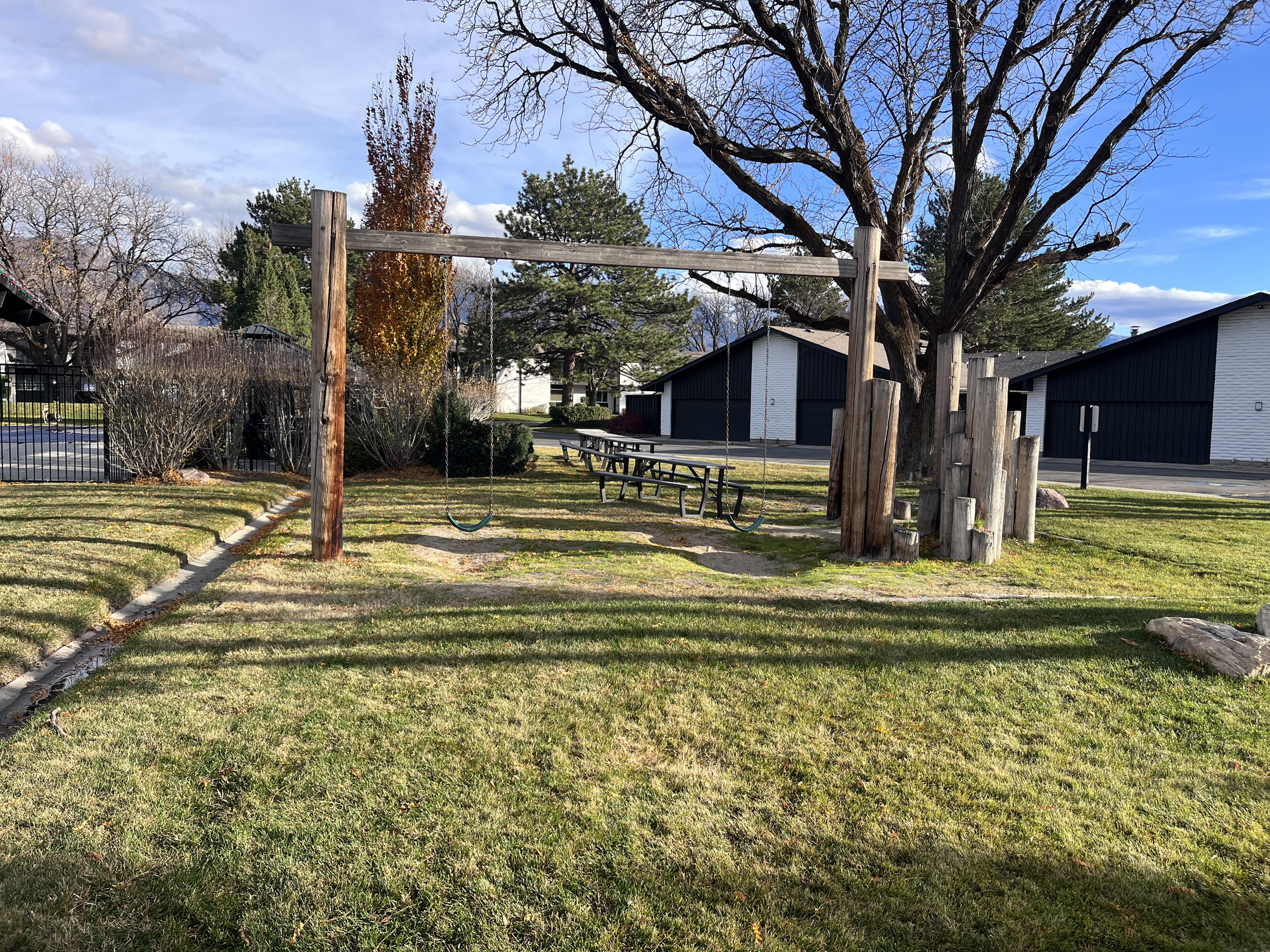 Community playground and grassed outdoor area featuring a swing set, picnic table, and mature trees