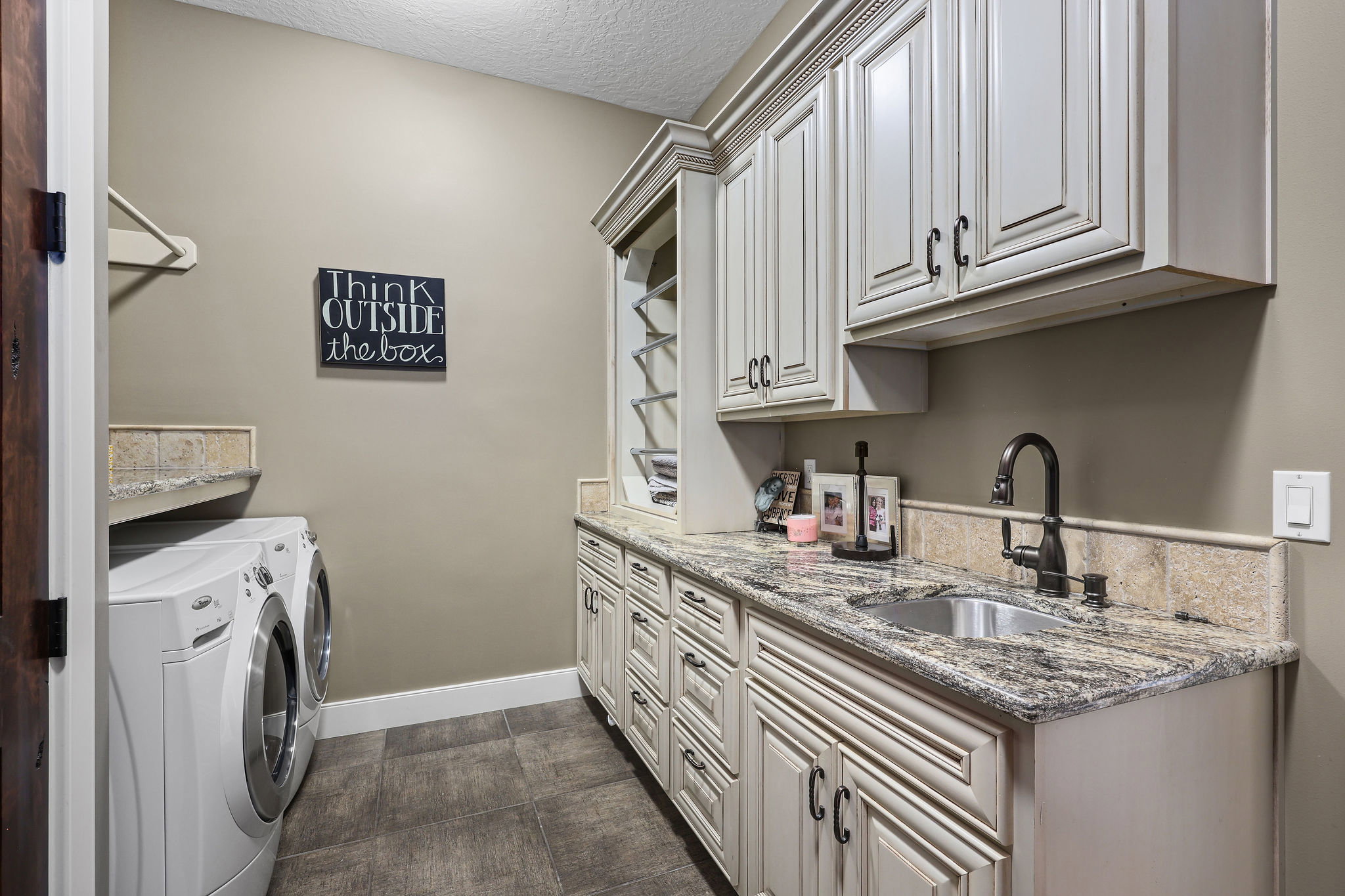 Laundry room featuring extensive custom cabinetry with distressed white finish, a stainless steel sink with a dark bronze faucet, and a stone countertop with a matching backsplash