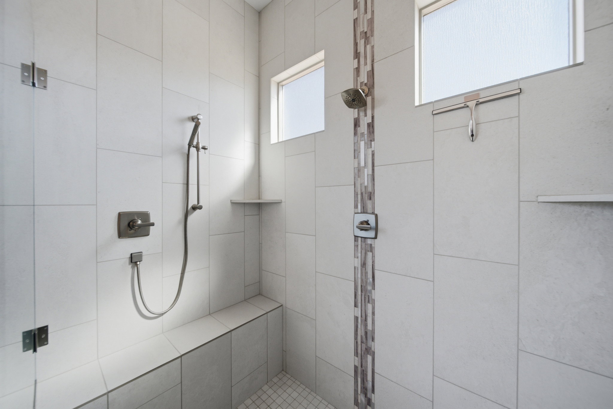 Spacious tiled shower featuring a built-in bench, dual recessed shelves, a handheld showerhead, a rainfall showerhead, and translucent horizontal windows