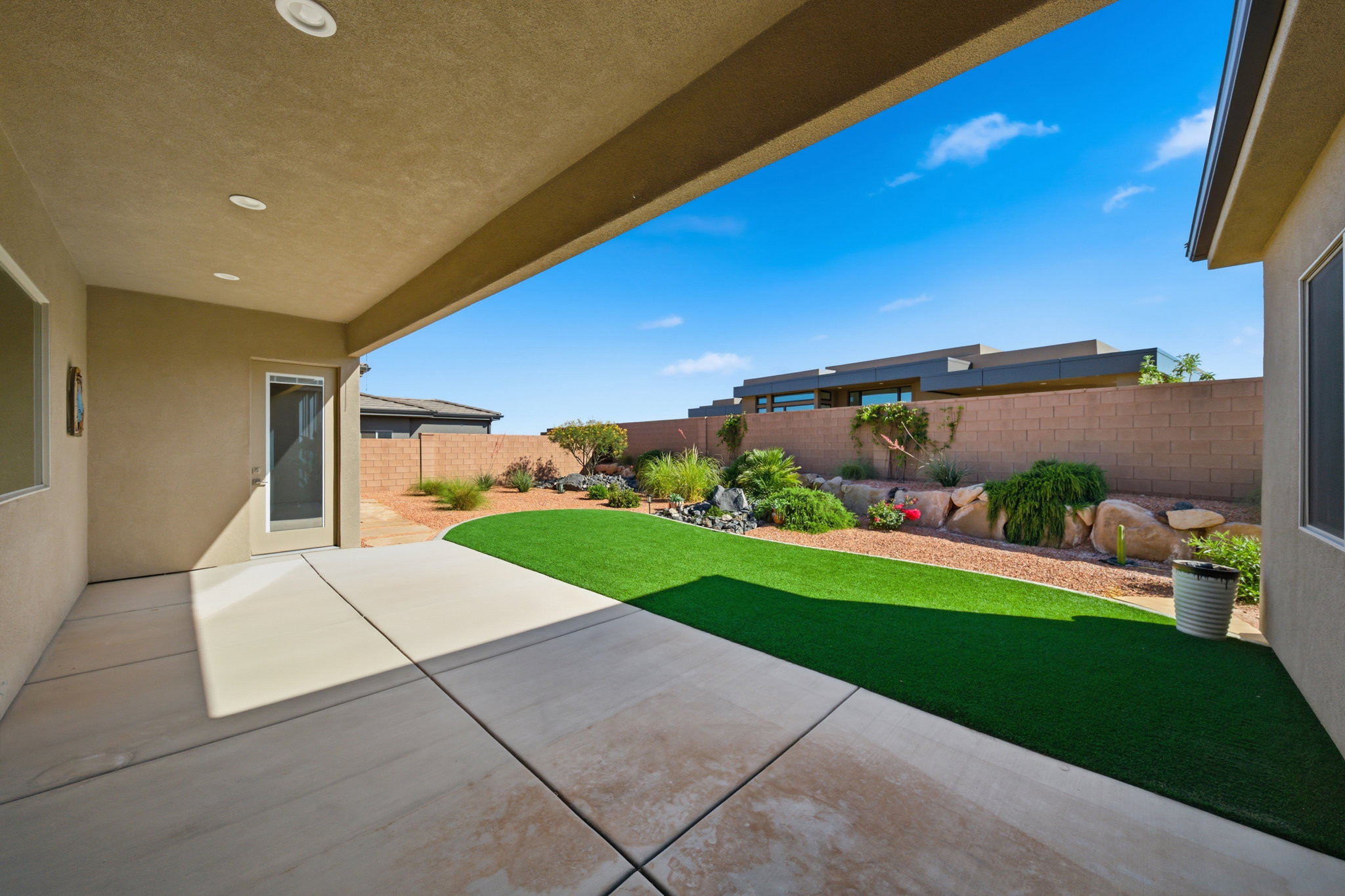 Covered patio with recessed lighting, concrete flooring, artificial turf lawn, and a stone-accented garden bed with drought-tolerant landscaping