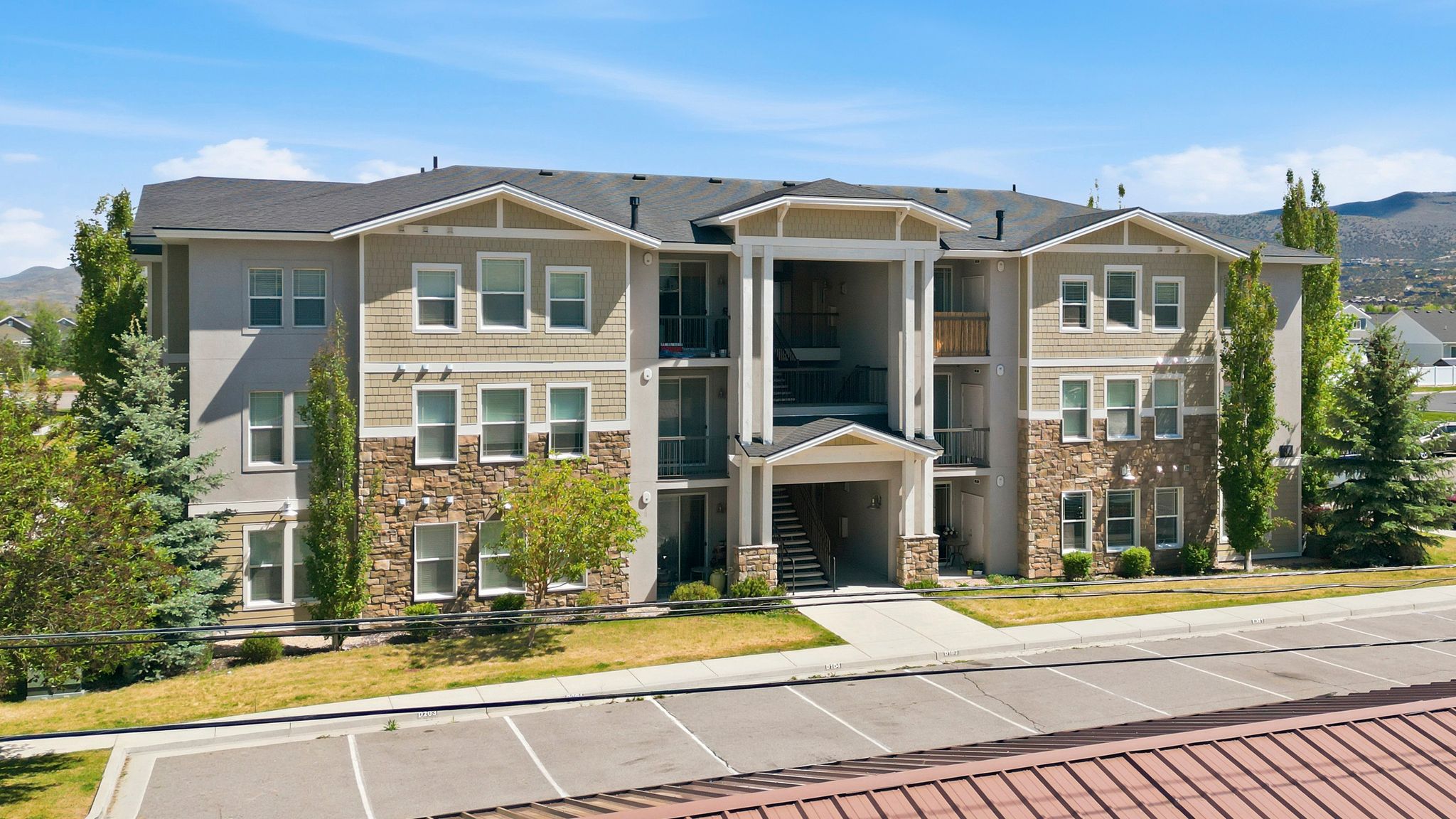 Multi-story building featuring a stone and siding facade, a covered entry with columns, private balconies, and a paved parking area