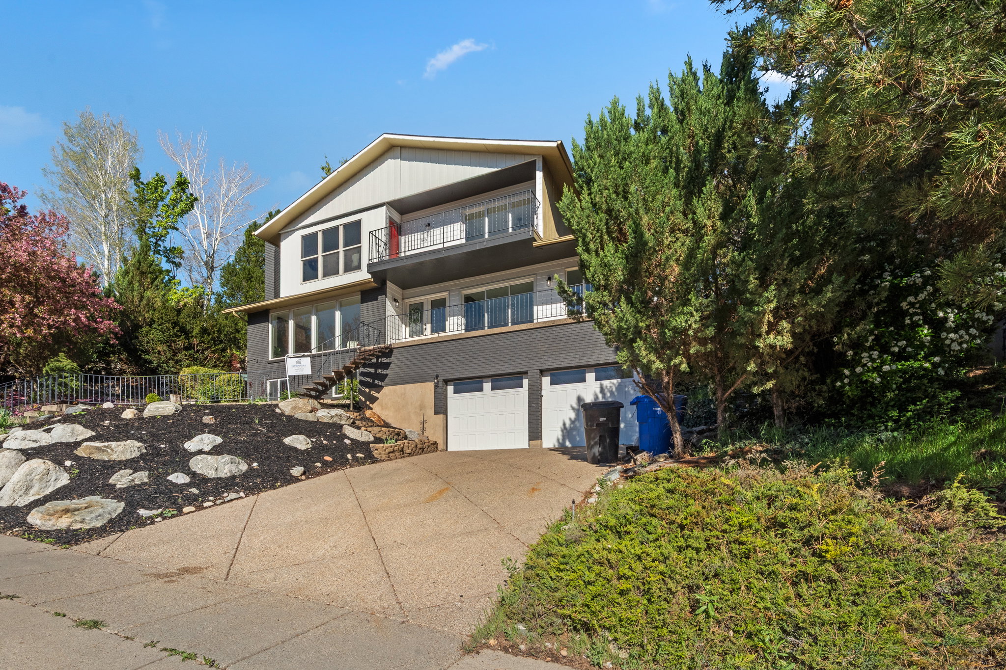 Multi-level residence featuring a two-car garage with white doors, a tiered front landscaping with rocks and dark ground cover, and mature trees