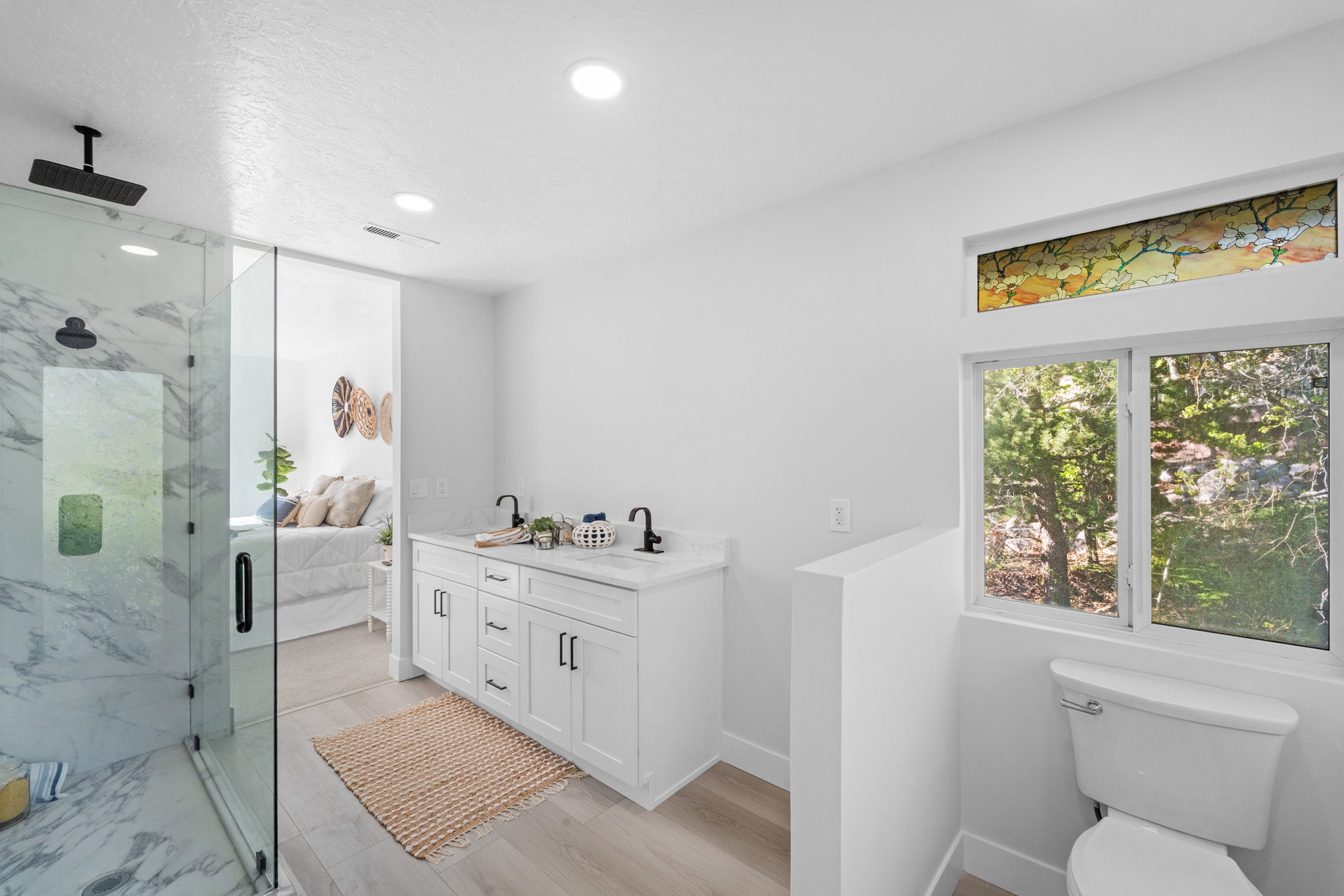 Spacious bathroom featuring a double vanity with white cabinetry, a glass-enclosed shower with marble-finish tiling, and a window with a decorative transom