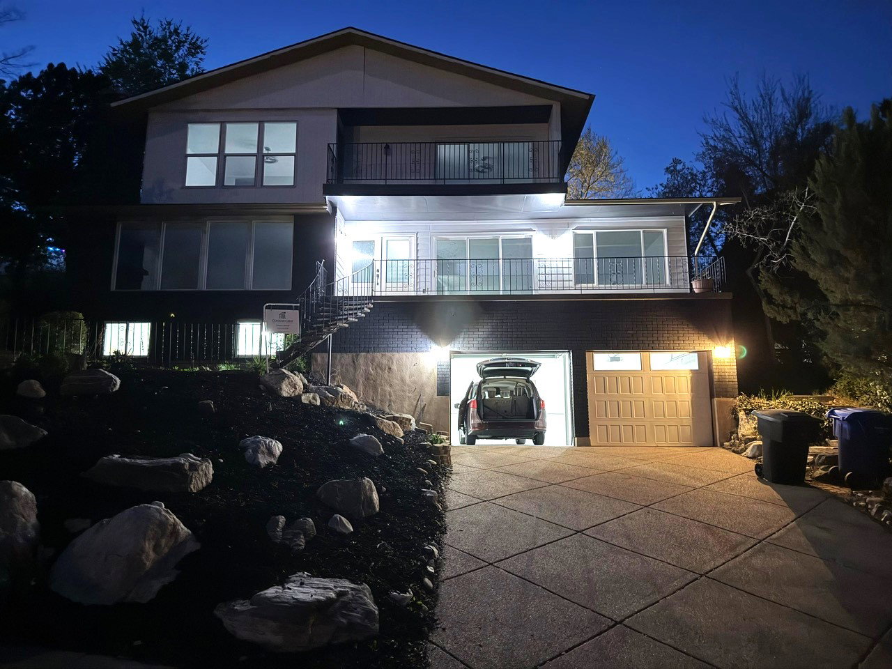 Multi-level exterior with a two-car garage, upper and lower balconies with black railings, and a landscaped rock garden