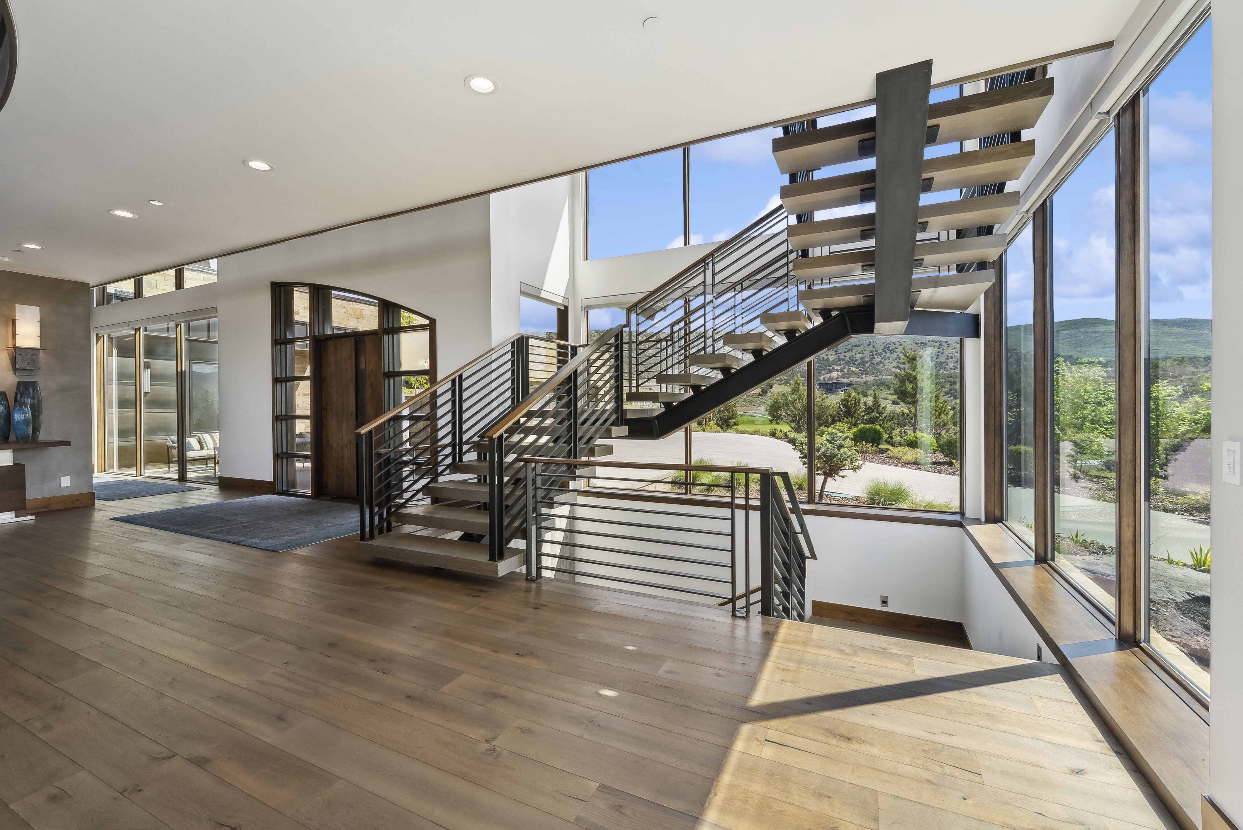 Floating staircase with open risers and metal railing, expansive window wall, wood-finish flooring, recessed lighting, and a modern entryway with glass paneling
