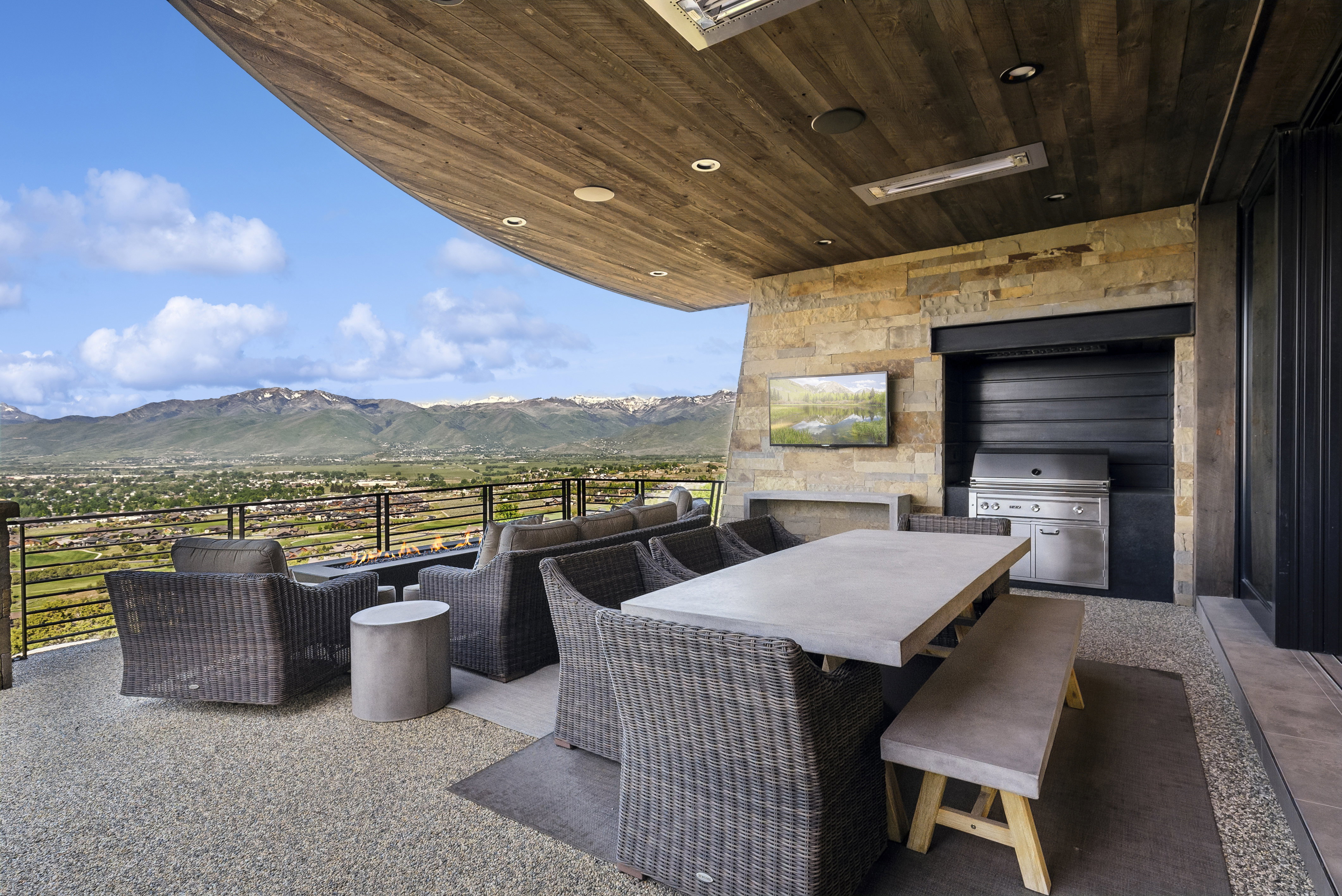 Spacious outdoor living area with a wood-finish ceiling, stone accent wall, integrated outdoor kitchen with a built-in grill, and a concrete dining table with bench seating