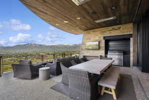 Spacious outdoor living area with a wood-finish ceiling, stone accent wall, integrated outdoor kitchen with a built-in grill, and a concrete dining table with bench seating