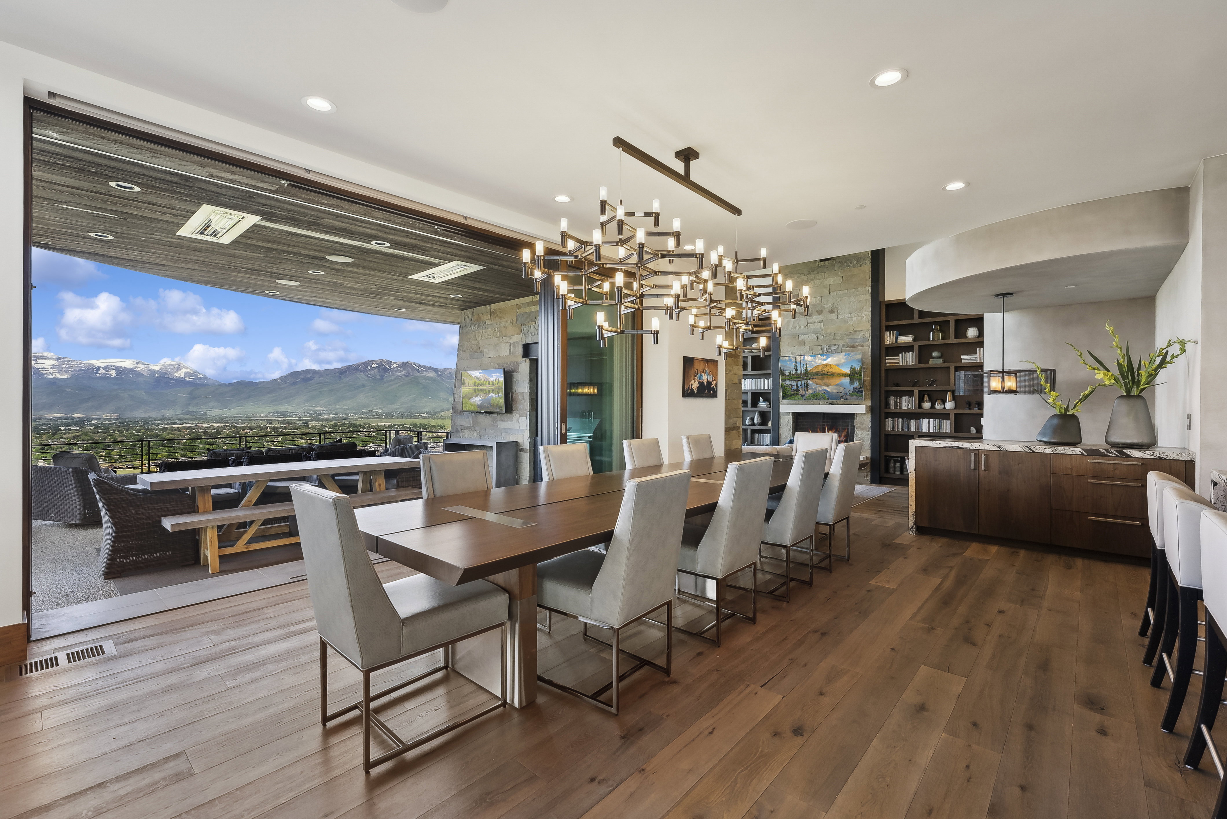 Expansive dining area featuring wood-finish flooring, a contemporary chandelier, and a retractable glass wall opening to a mountain vista