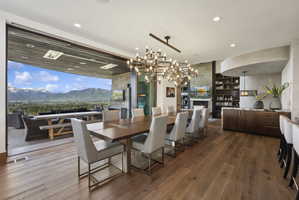 Expansive dining area featuring wood-finish flooring, a contemporary chandelier, and a retractable glass wall opening to a mountain vista