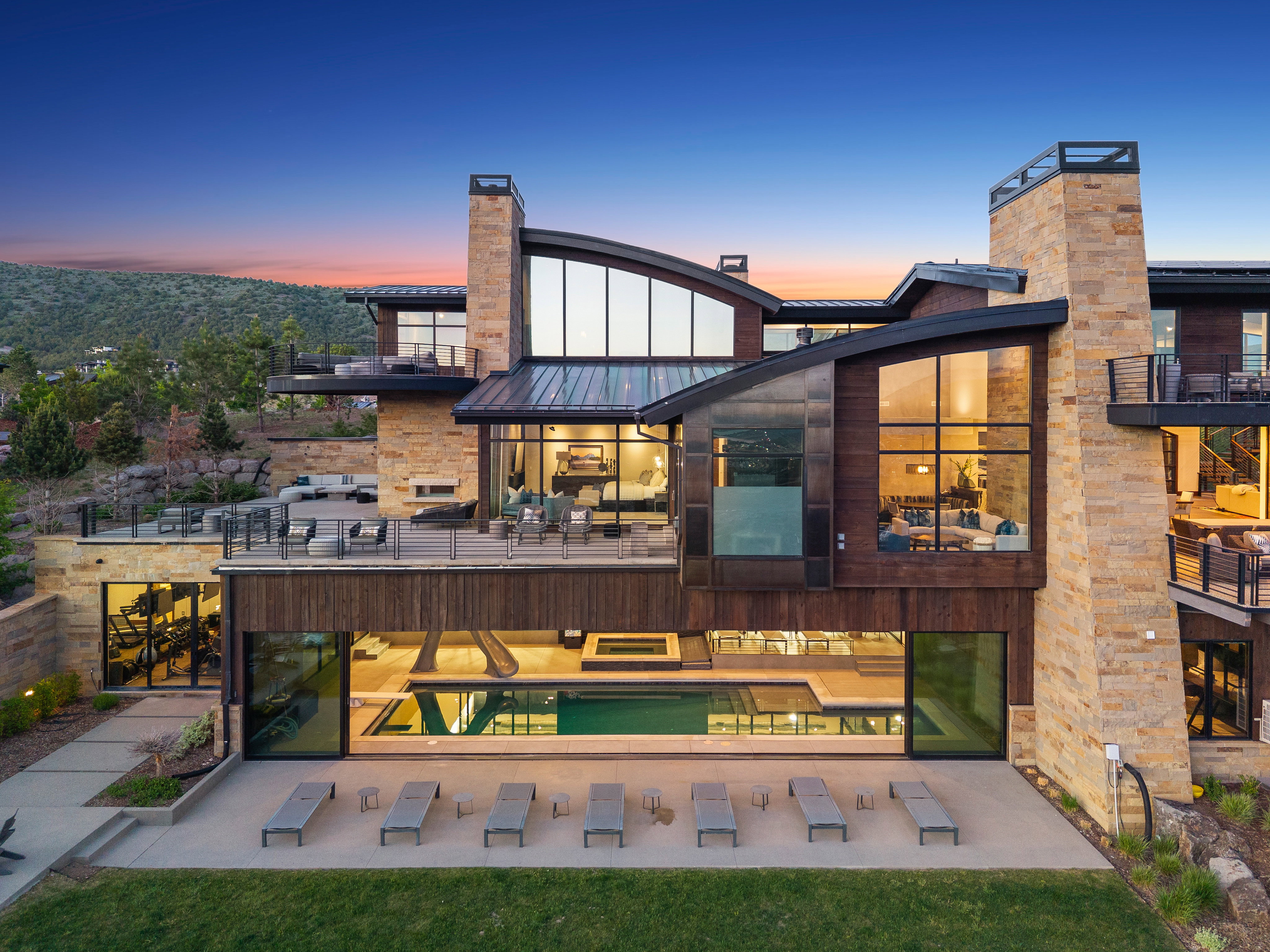 Contemporary multi-level residence featuring stone and wood-finish siding, extensive window walls, multiple outdoor terraces, and an indoor swimming pool with an adjacent patio
