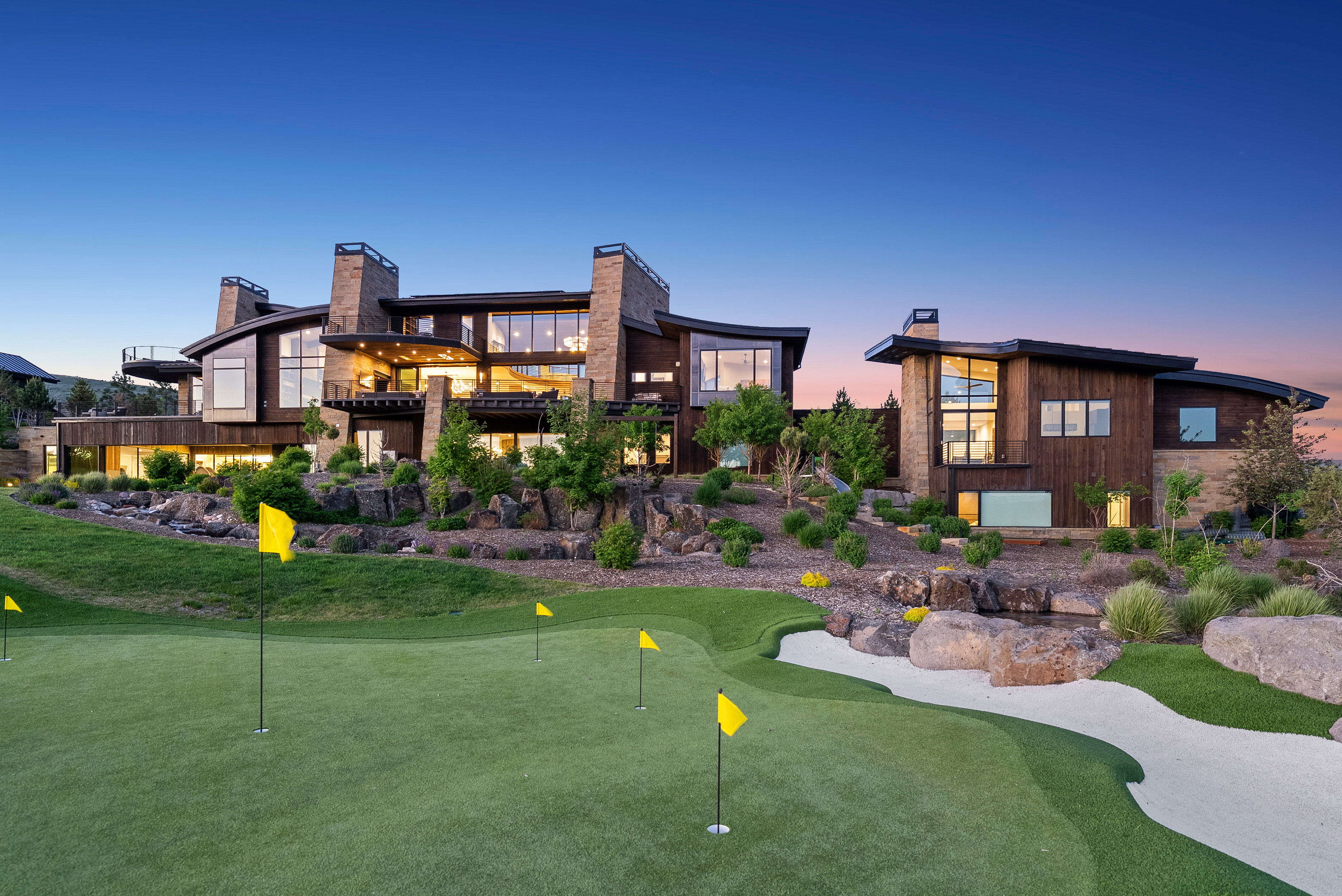 Expansive putting green with sand trap, set against a multi-level residence featuring extensive glazing, stone accents, wood siding, and multiple outdoor balconies