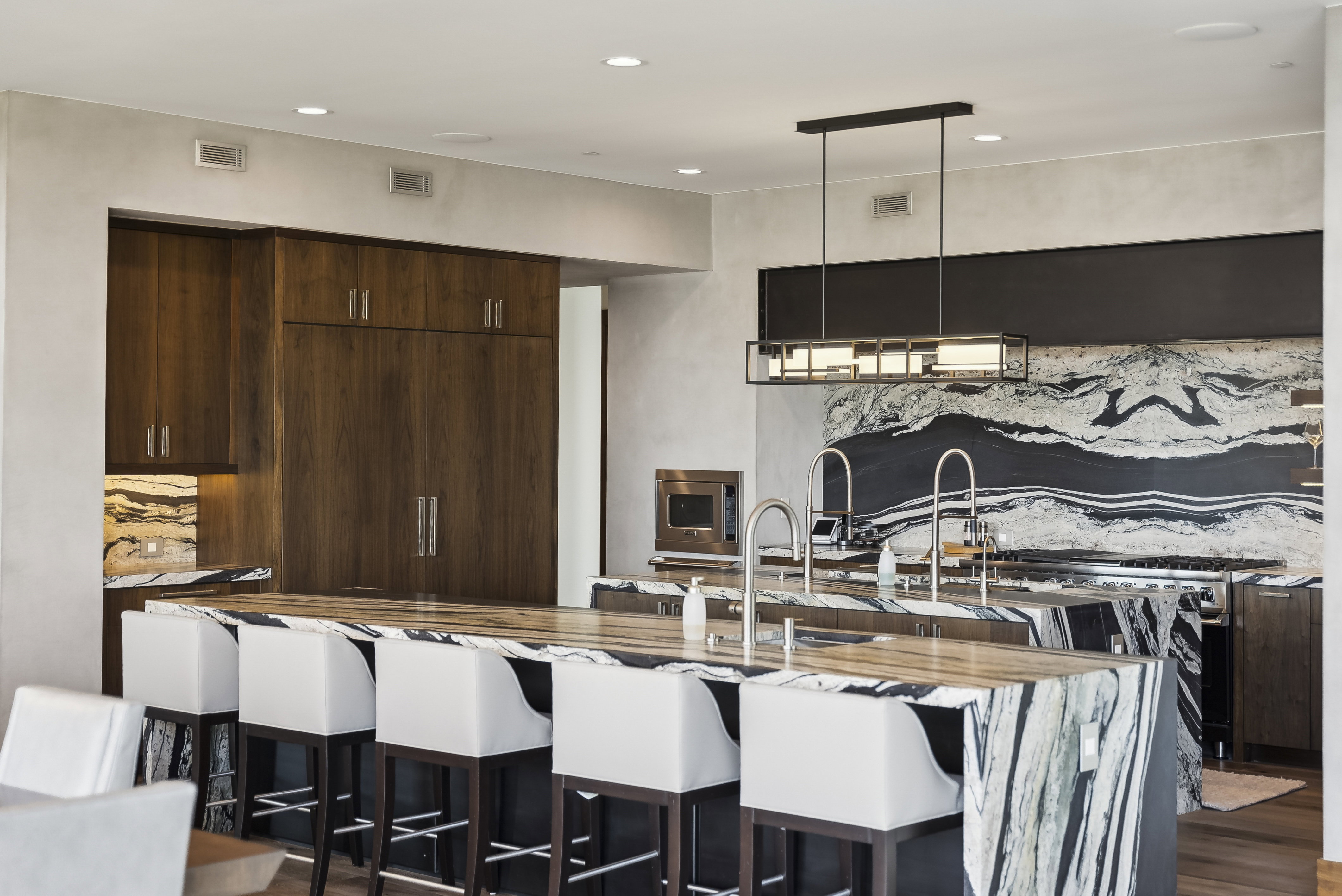 Kitchen featuring an expansive island with waterfall-edge stone countertop, wood-finish cabinetry, built-in stainless steel appliances, a modern linear pendant light fixture, and a dramatic stone slab backsplash