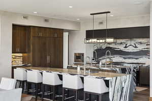 Kitchen featuring an expansive island with waterfall-edge stone countertop, wood-finish cabinetry, built-in stainless steel appliances, a modern linear pendant light fixture, and a dramatic stone slab backsplash