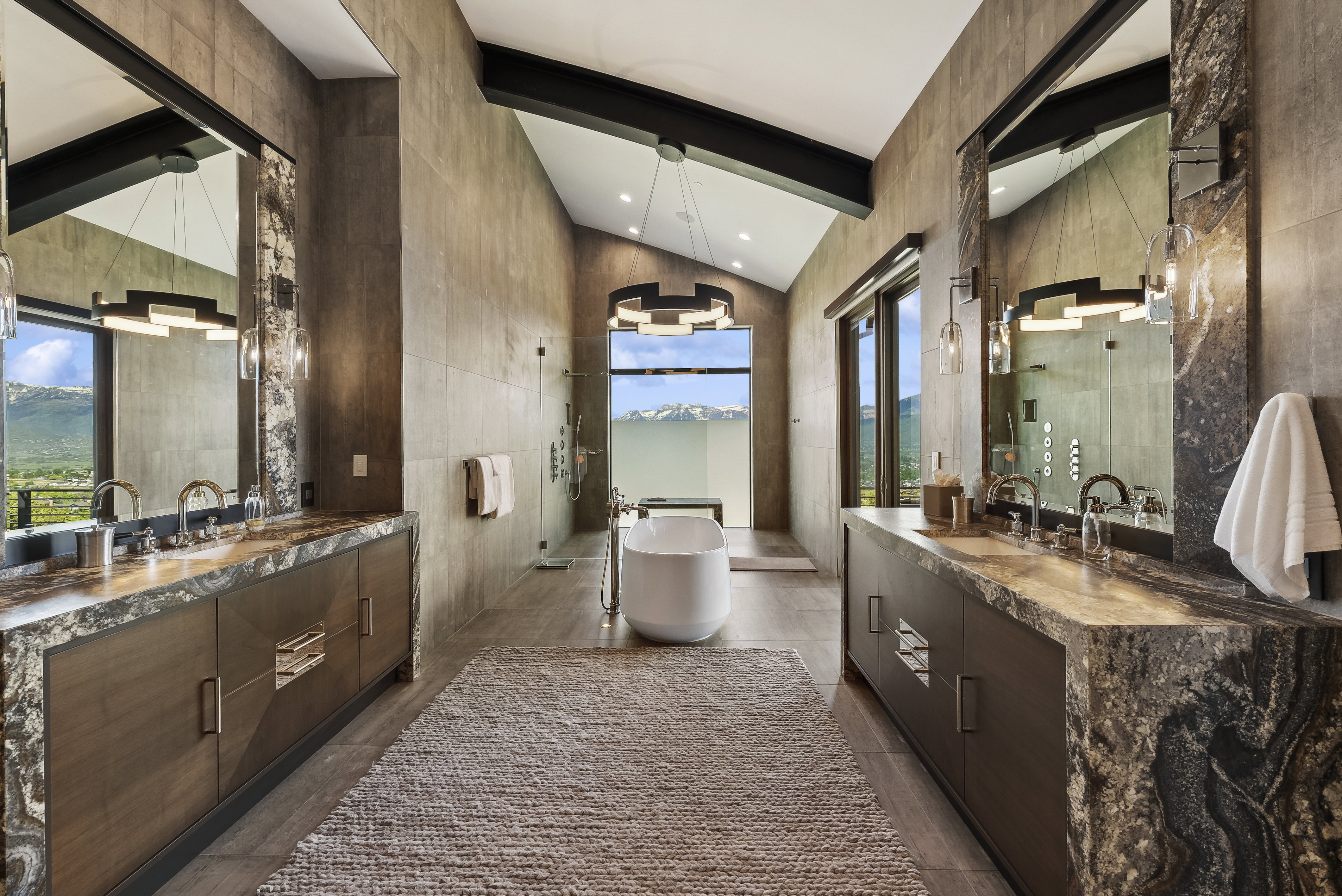 Vaulted ceiling bathroom featuring a freestanding tub, expansive windows, dual vanities with stone countertops, and a glass-enclosed shower