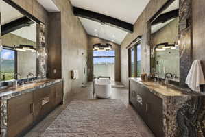 Vaulted ceiling bathroom featuring a freestanding tub, expansive windows, dual vanities with stone countertops, and a glass-enclosed shower