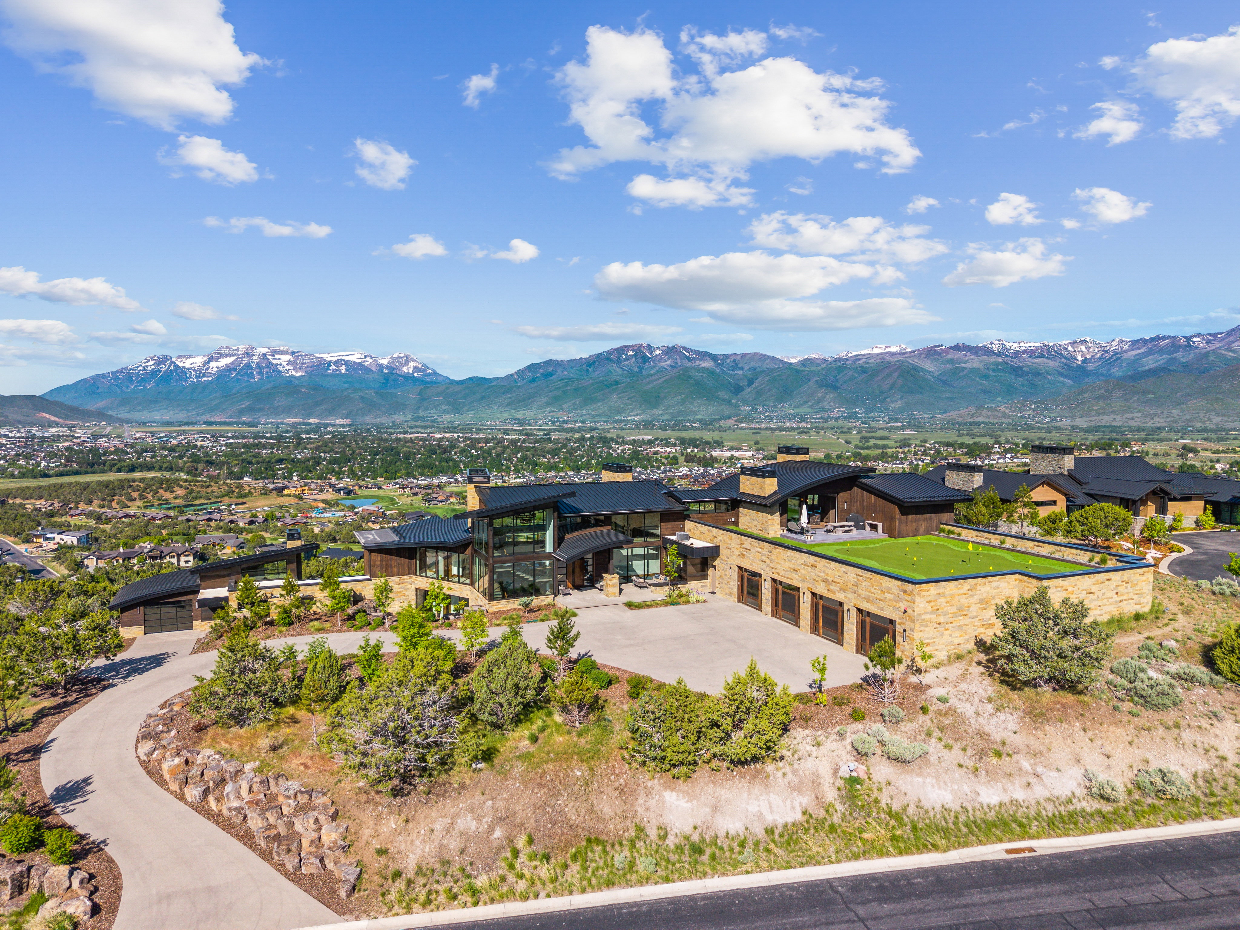 Expansive contemporary estate featuring stone and wood facade, a green roof putting area, multiple garages, and a paved driveway with natural rock landscaping