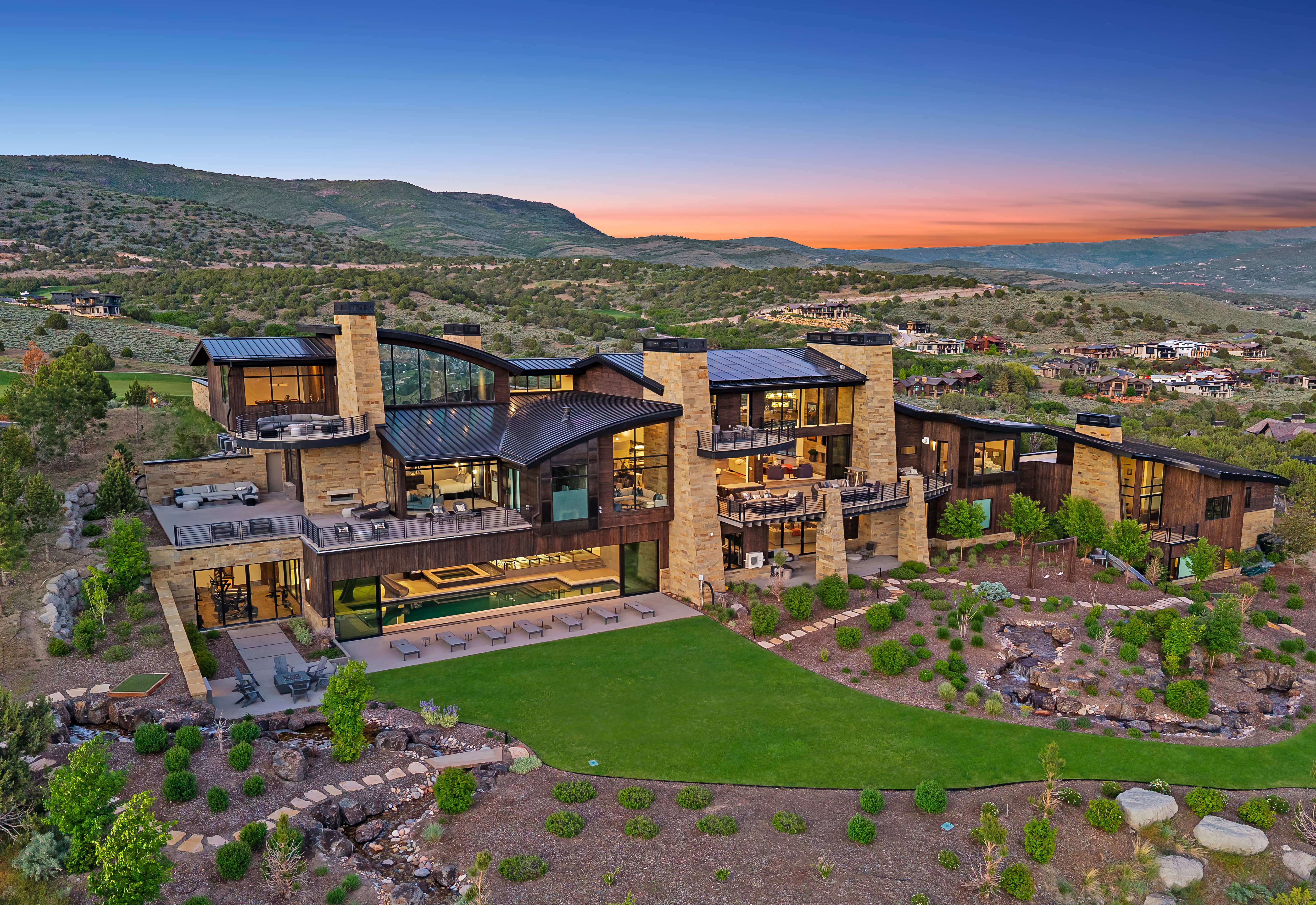 Expansive contemporary residence featuring stone and wood-panel exterior finishes, multiple balconies, a lower-level patio with an integrated pool, and a manicured lawn with natural rock landscaping