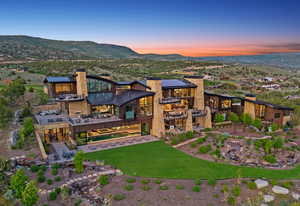 Expansive contemporary residence featuring stone and wood-panel exterior finishes, multiple balconies, a lower-level patio with an integrated pool, and a manicured lawn with natural rock landscaping