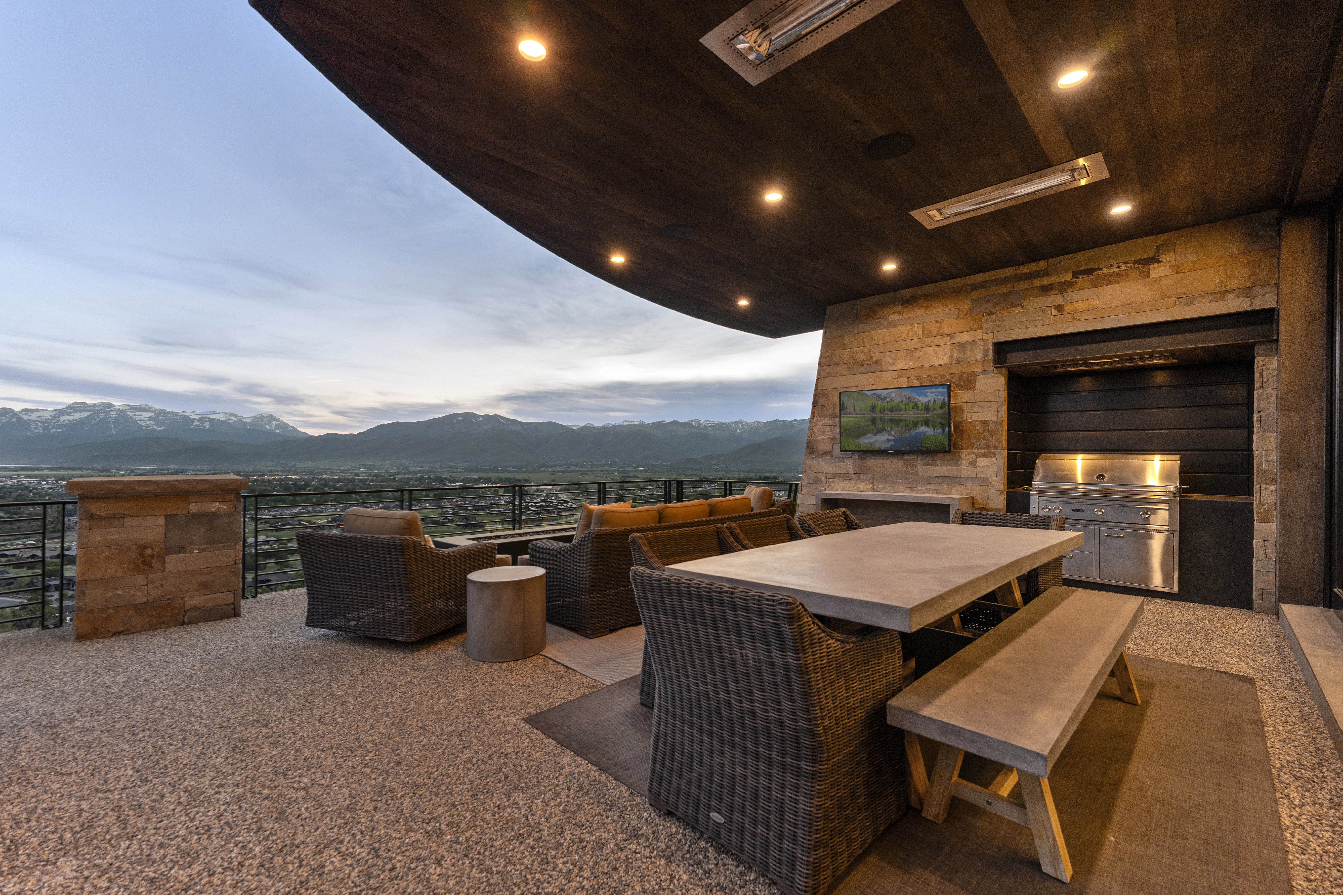 Expansive covered patio featuring an exposed aggregate floor, stone wall accents, a built-in outdoor kitchen with a stainless steel grill, a mounted television, and recessed ceiling lighting