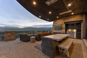 Expansive covered patio featuring an exposed aggregate floor, stone wall accents, a built-in outdoor kitchen with a stainless steel grill, a mounted television, and recessed ceiling lighting