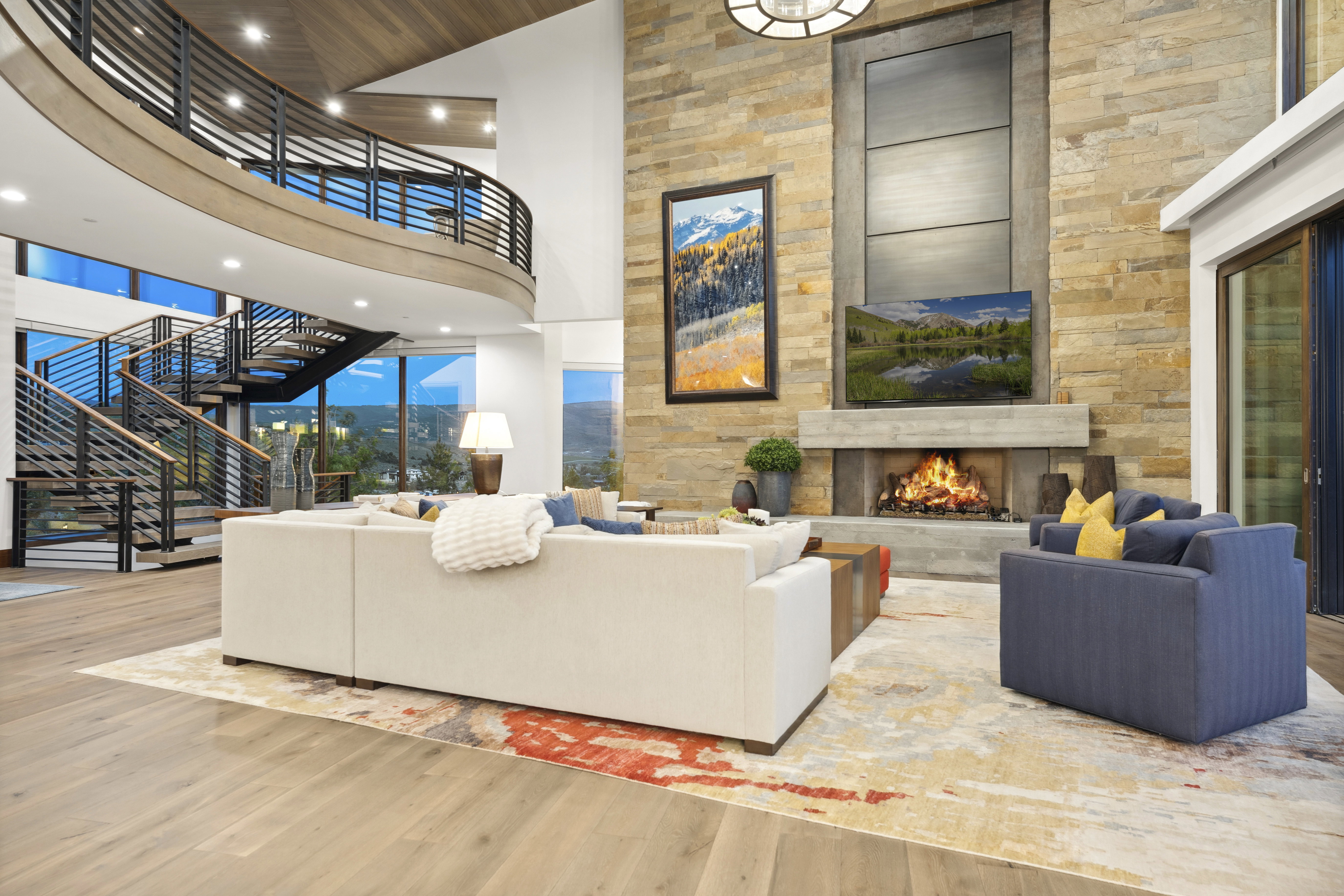Spacious living area featuring a floor-to-ceiling stacked stone fireplace with a concrete mantel, wood-finish flooring, and a multi-level open-tread staircase with metal railings