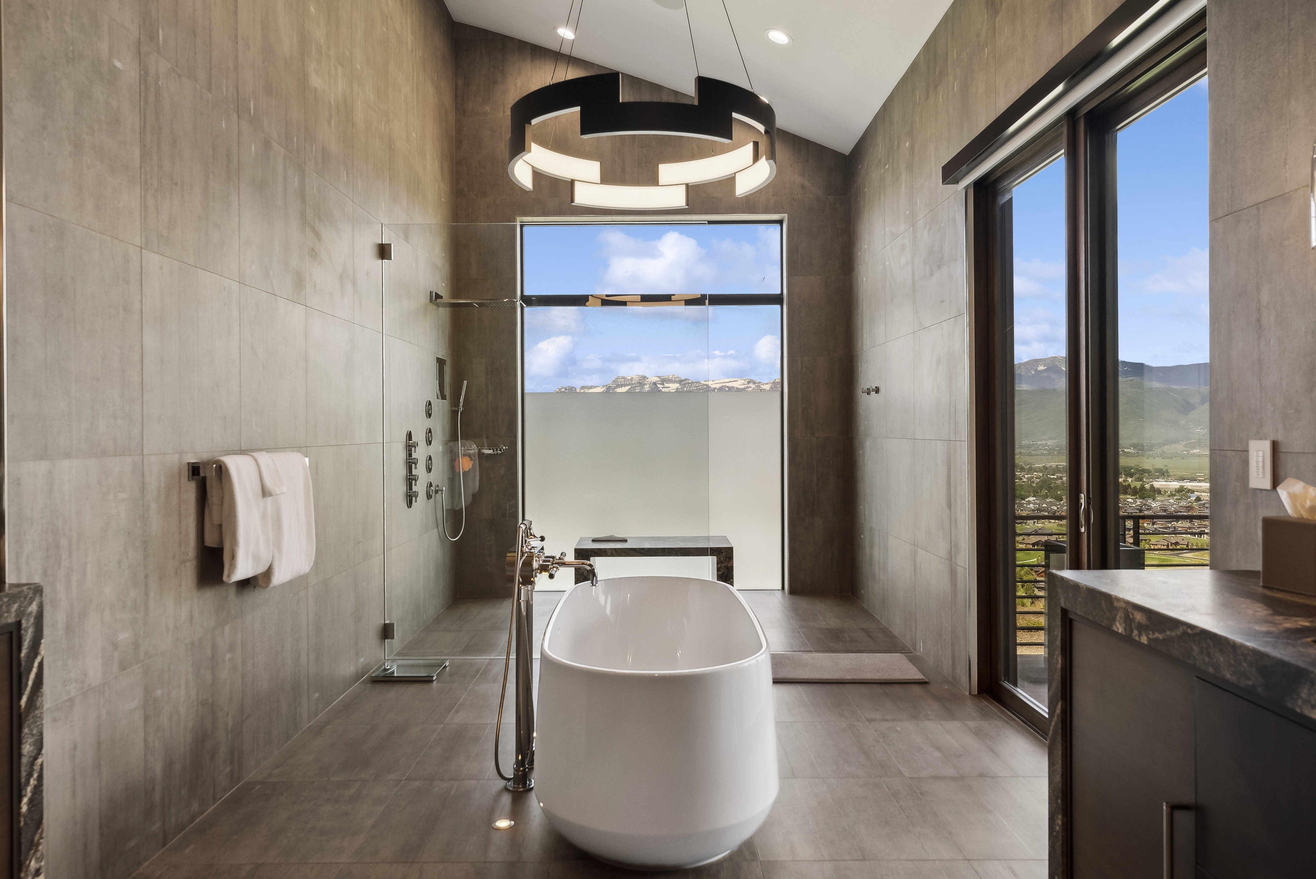 Freestanding soaking tub with floor-mounted faucet, oversized tile flooring, and a glass-enclosed shower featuring a wall-mounted panel