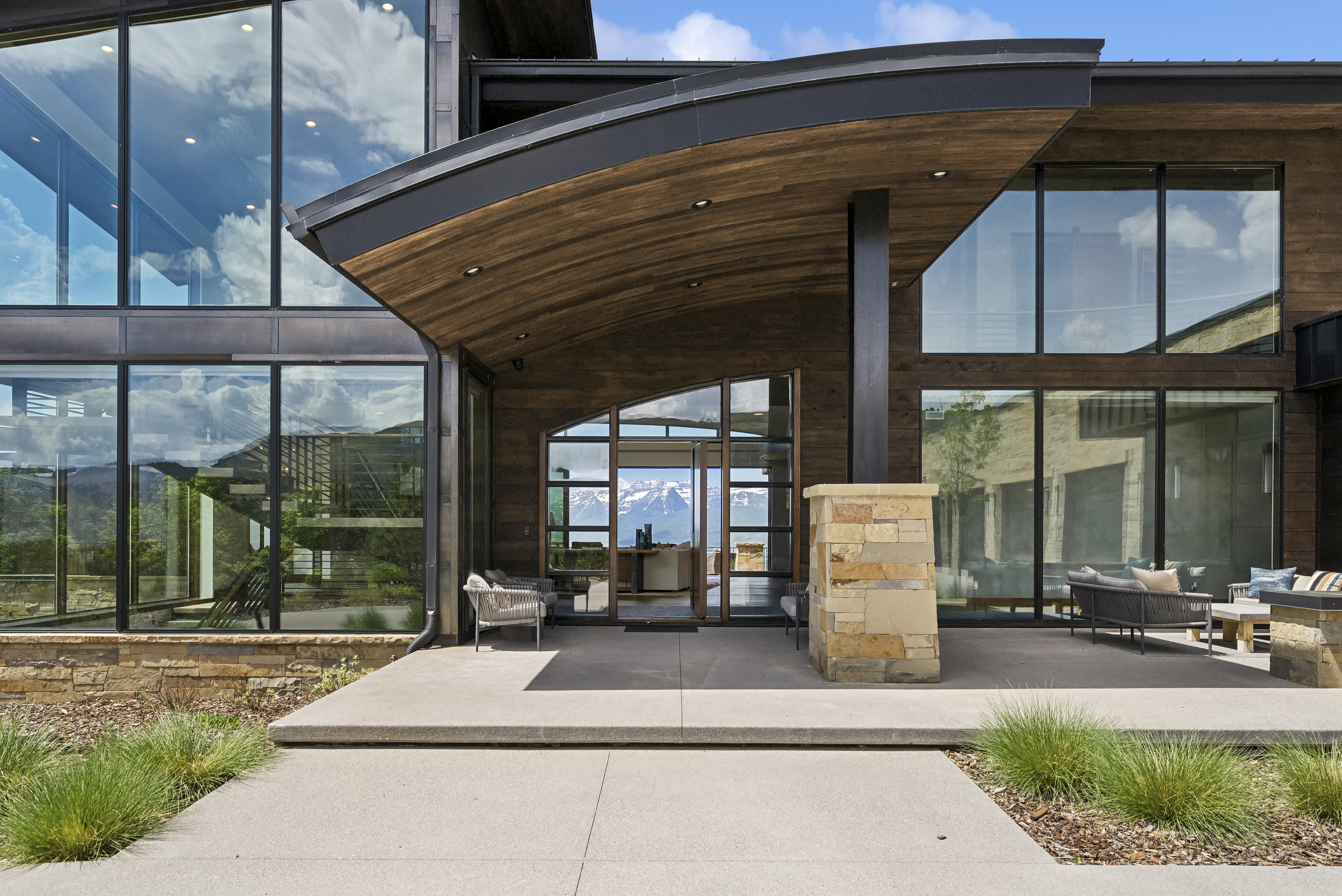 Contemporary architectural design featuring extensive window walls, wood-paneled exterior accents, a curved portico, and a stone-clad column