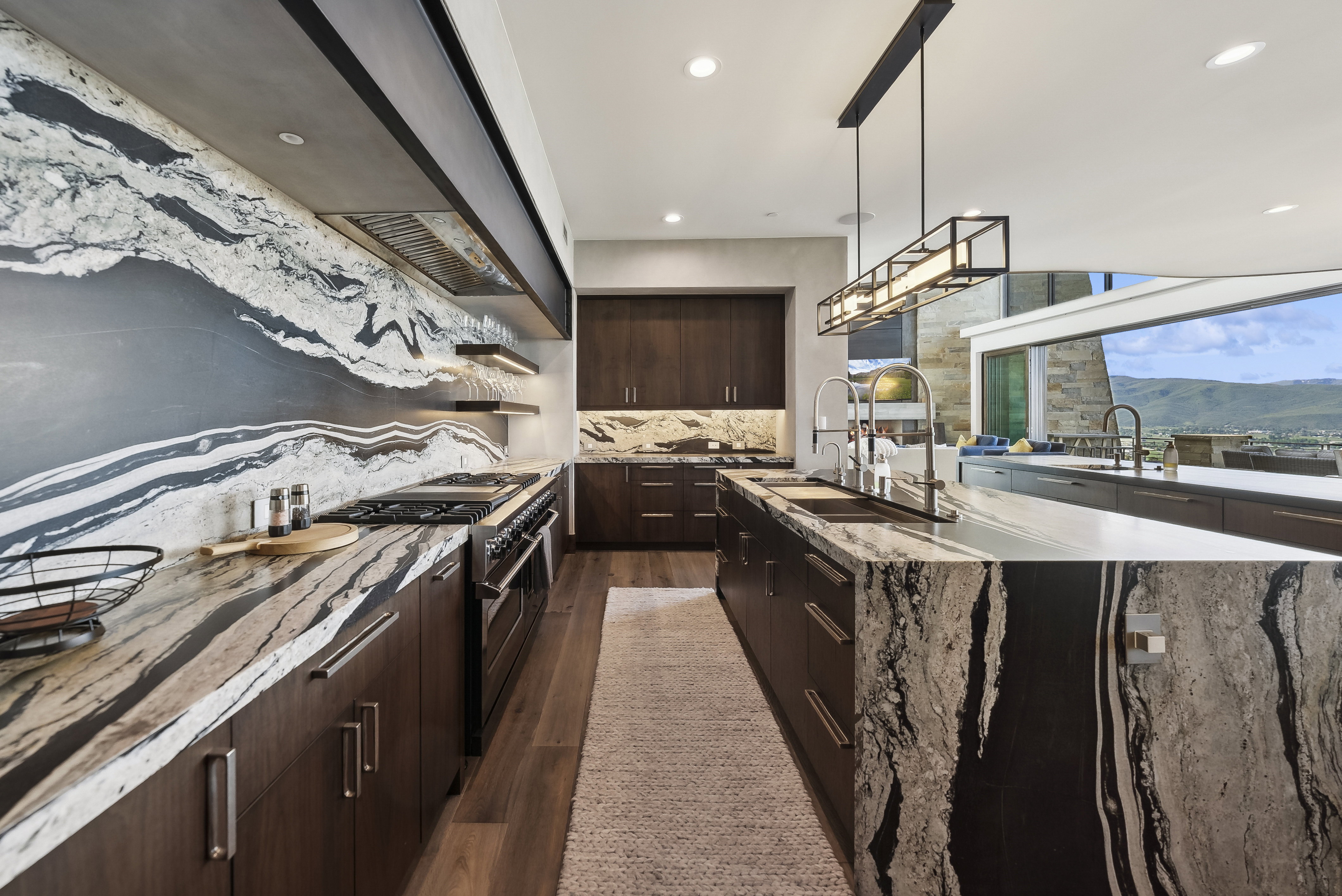 Gourmet kitchen featuring a waterfall island, custom dark wood cabinetry, a professional-grade range, integrated range hood, and a dramatic stone backsplash