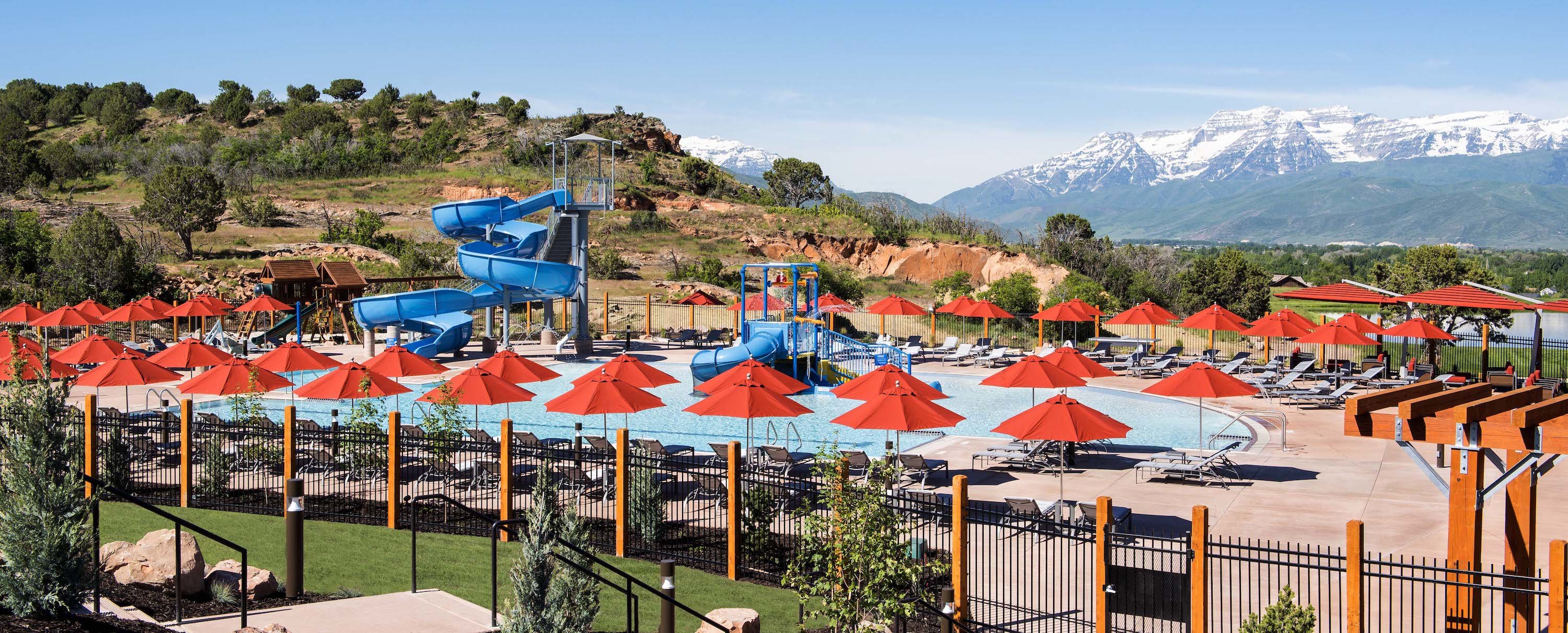 Resort-style pool area featuring multiple blue water slides, numerous red umbrellas, and lounge seating