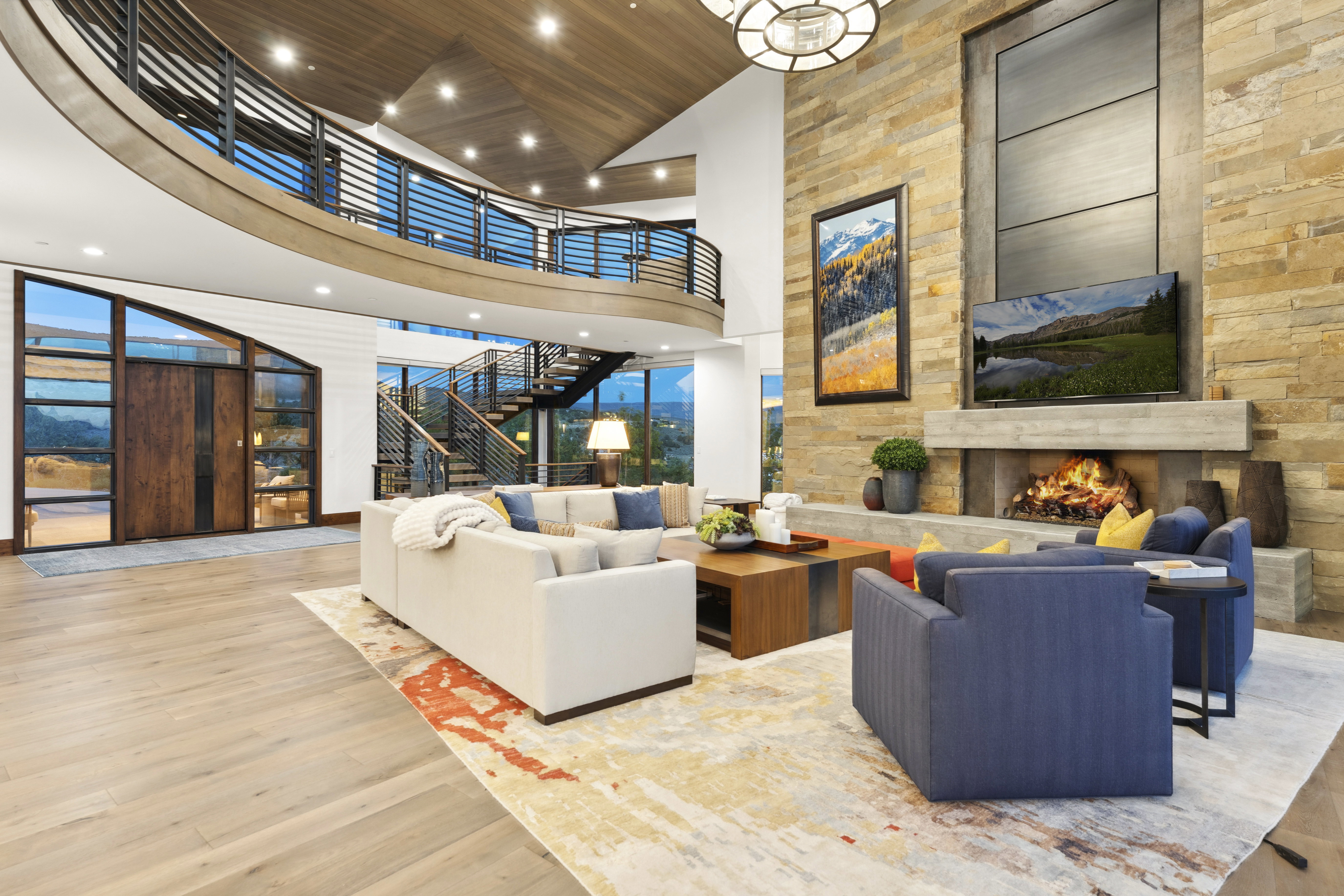 Grand living space featuring a two-story stone fireplace with a substantial hearth, wood-finish flooring, and a curved second-story balcony with metal railings