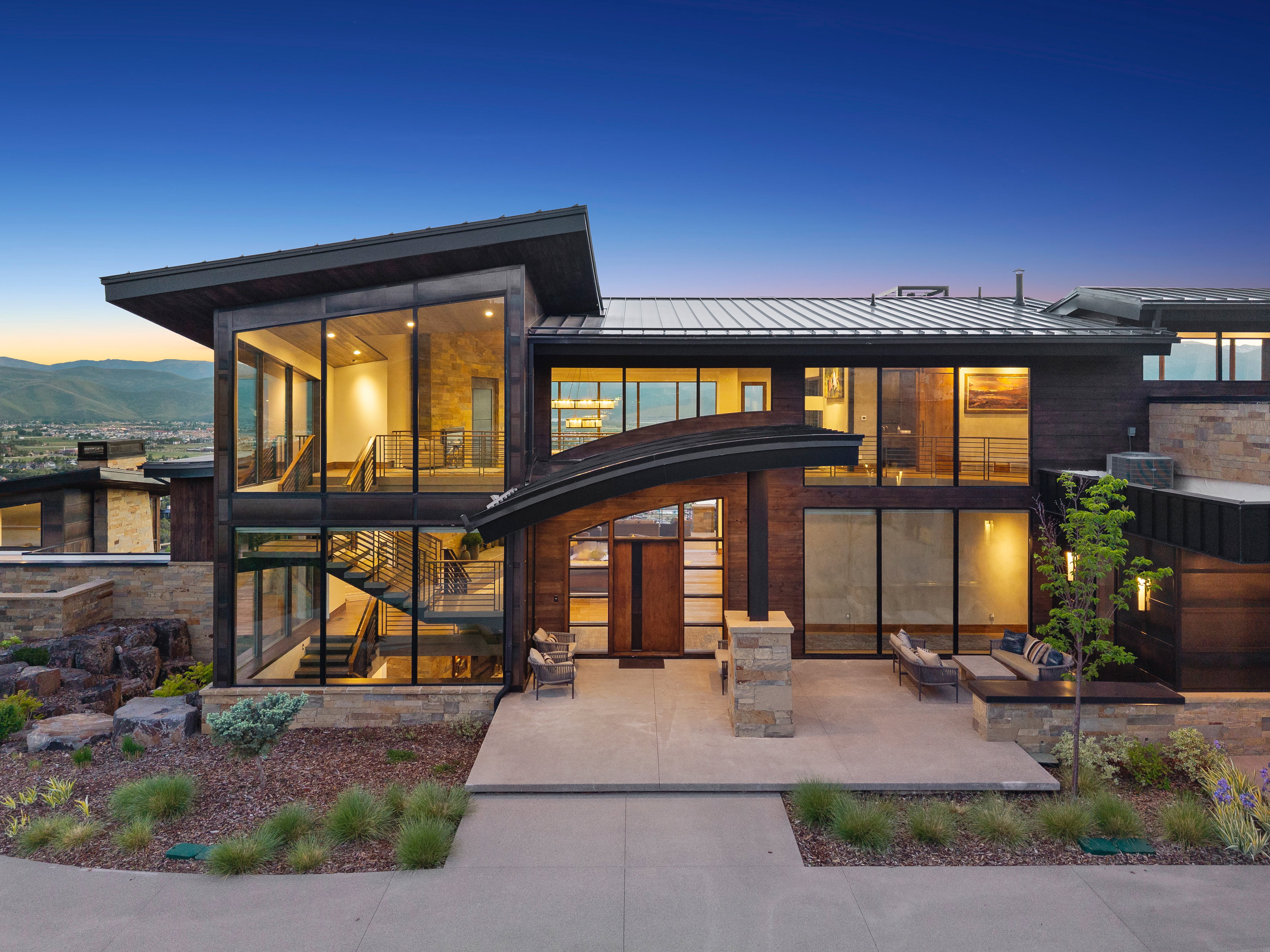 Contemporary architectural design featuring extensive window walls, a metal roof, stacked stone accents, and a prominent wood-finish entry door
