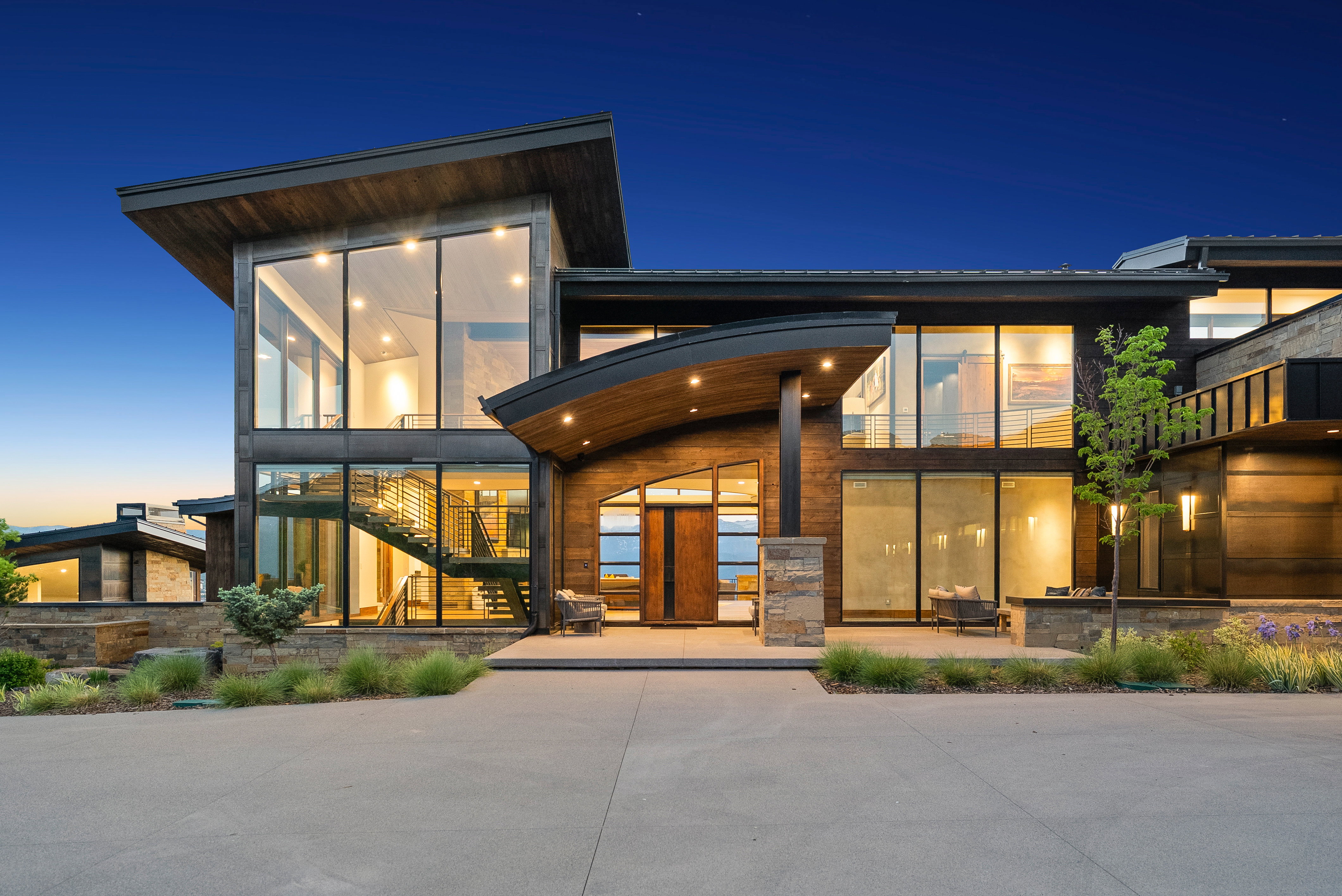 Contemporary architectural design featuring expansive glass panels, wood siding, stone accents, and a curved entryway canopy