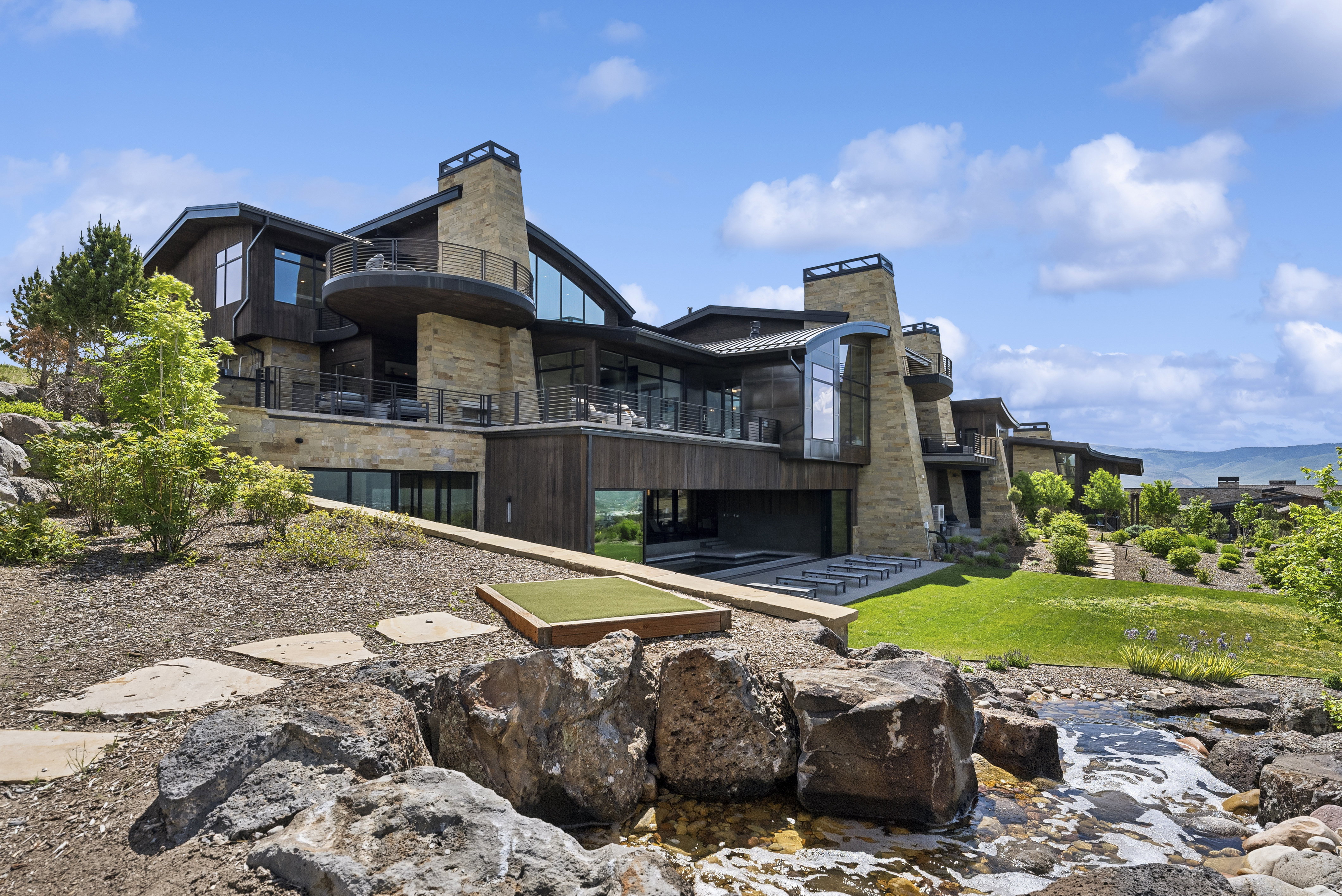 Multi-level modern home featuring stone and wood-finish siding, extensive glass, multiple balconies, and a putting green
