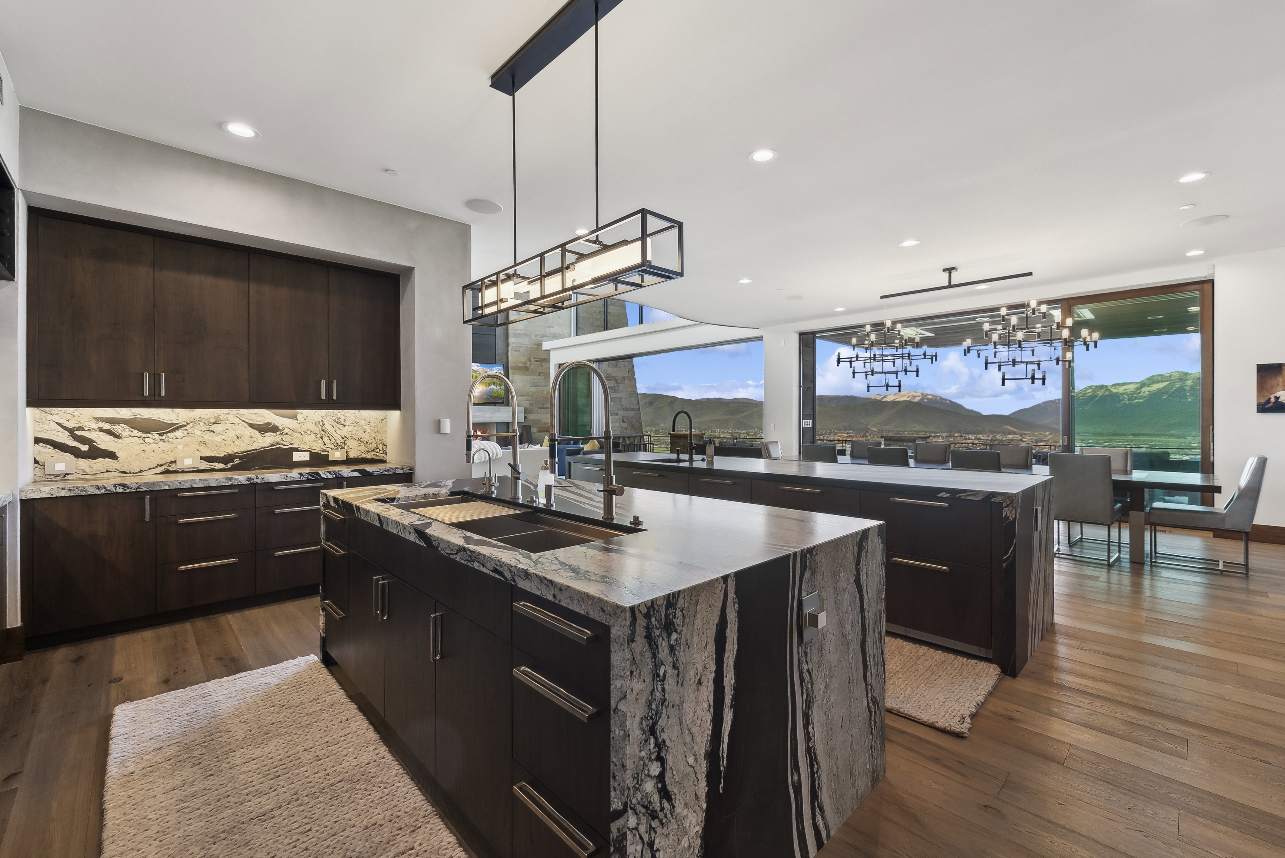 Expansive kitchen featuring dual waterfall islands with integrated sinks and dark cabinetry, complemented by a light-colored stone backsplash with under-cabinet lighting
