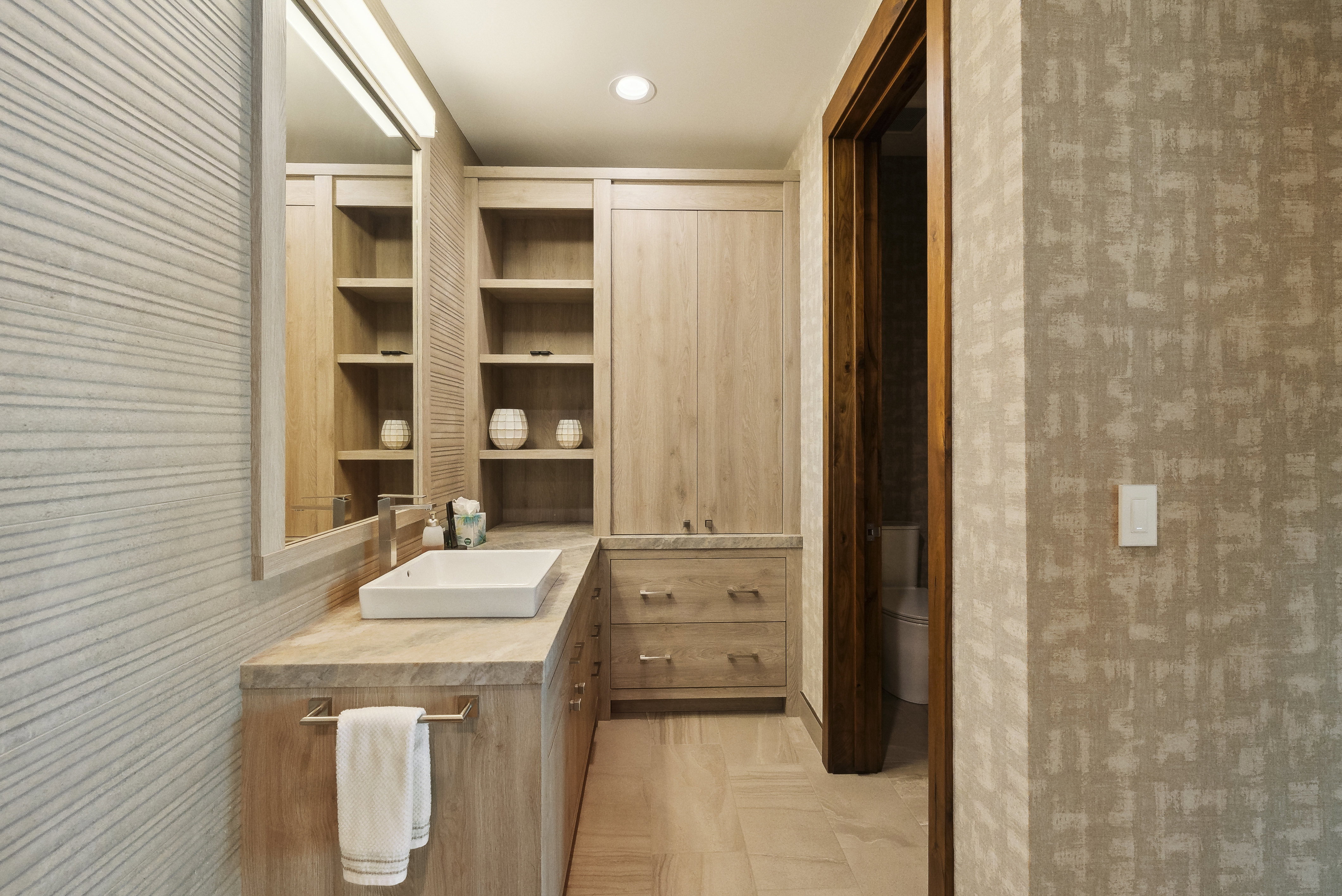 Bathroom with a rectangular vessel sink, integrated wood-finish cabinetry, built-in shelving, wood-finish countertop, and patterned wall covering