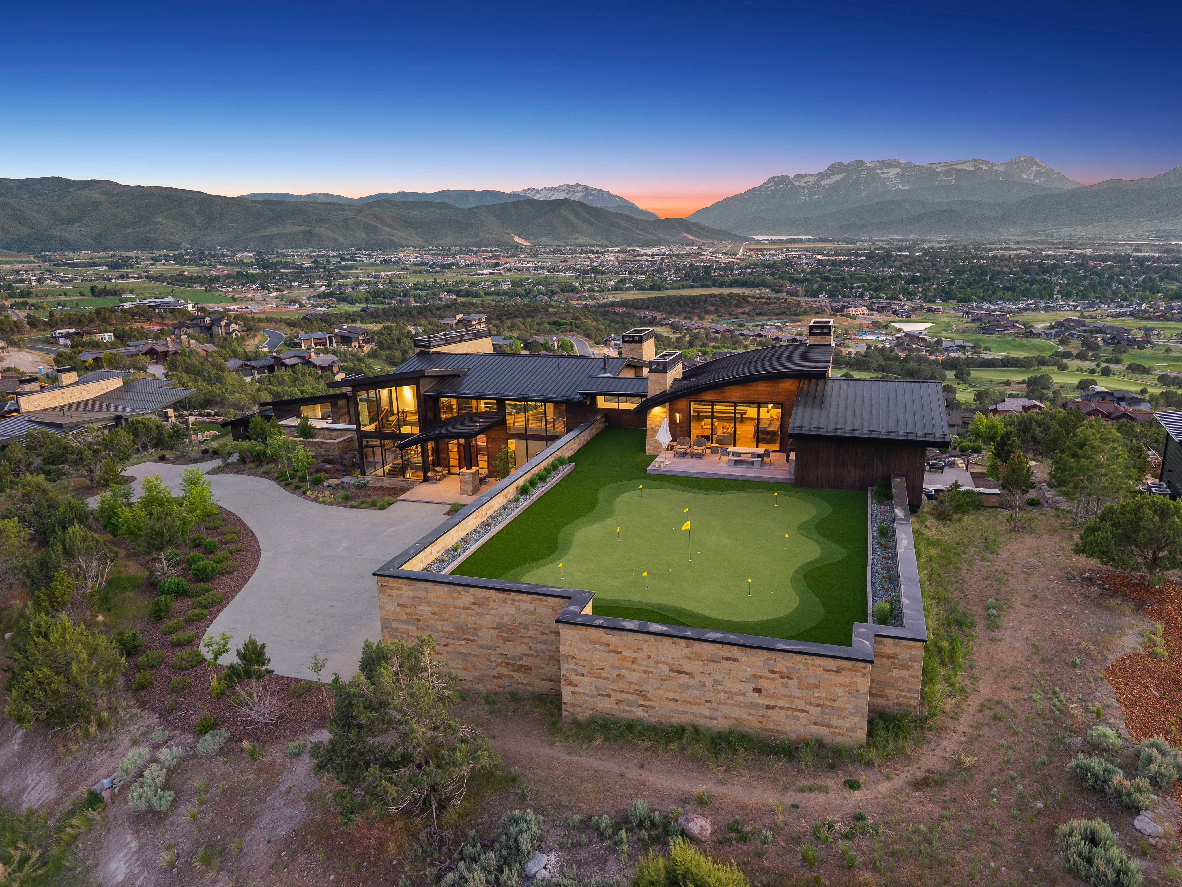 Modern architectural residence featuring extensive glass facades, a large putting green, a stone retaining wall, and a curved driveway