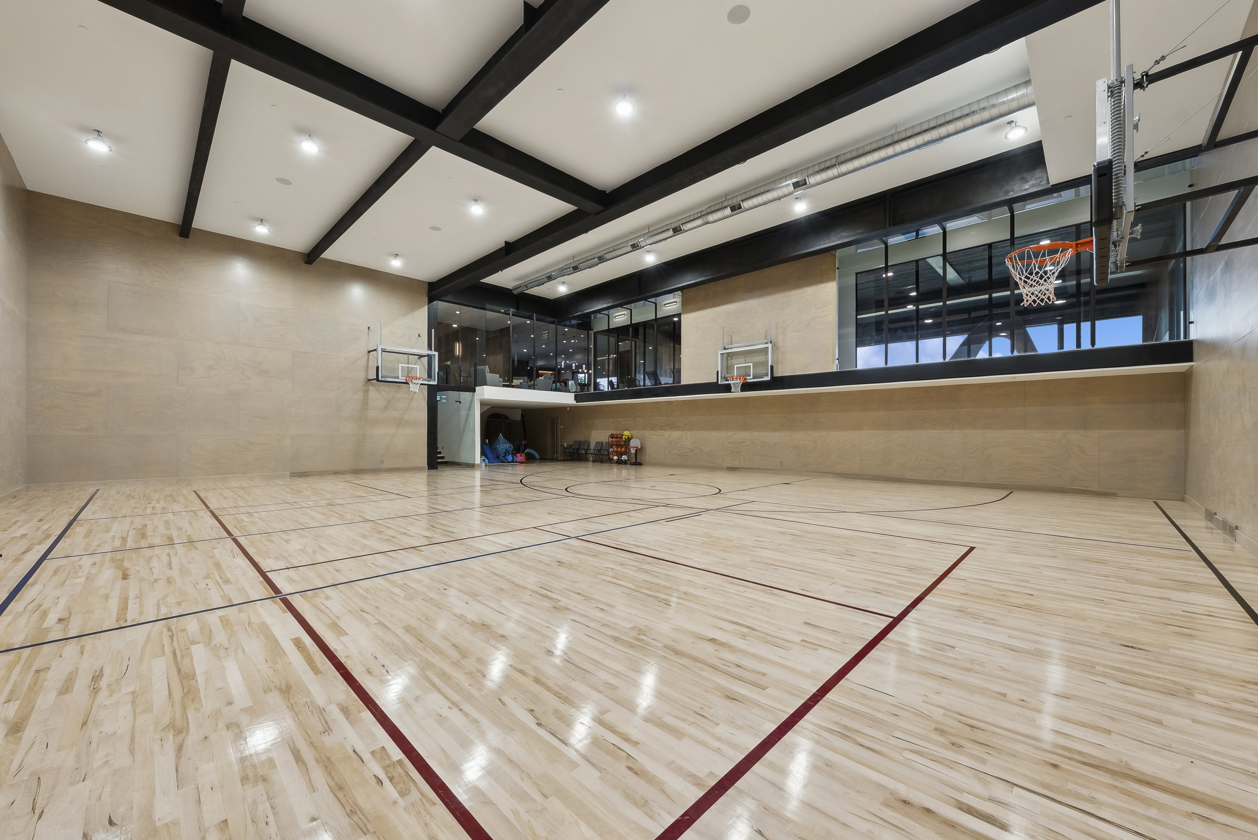 Indoor multi-sport court featuring light wood-finish flooring with marked lines, multiple basketball hoops, and recessed ceiling lighting