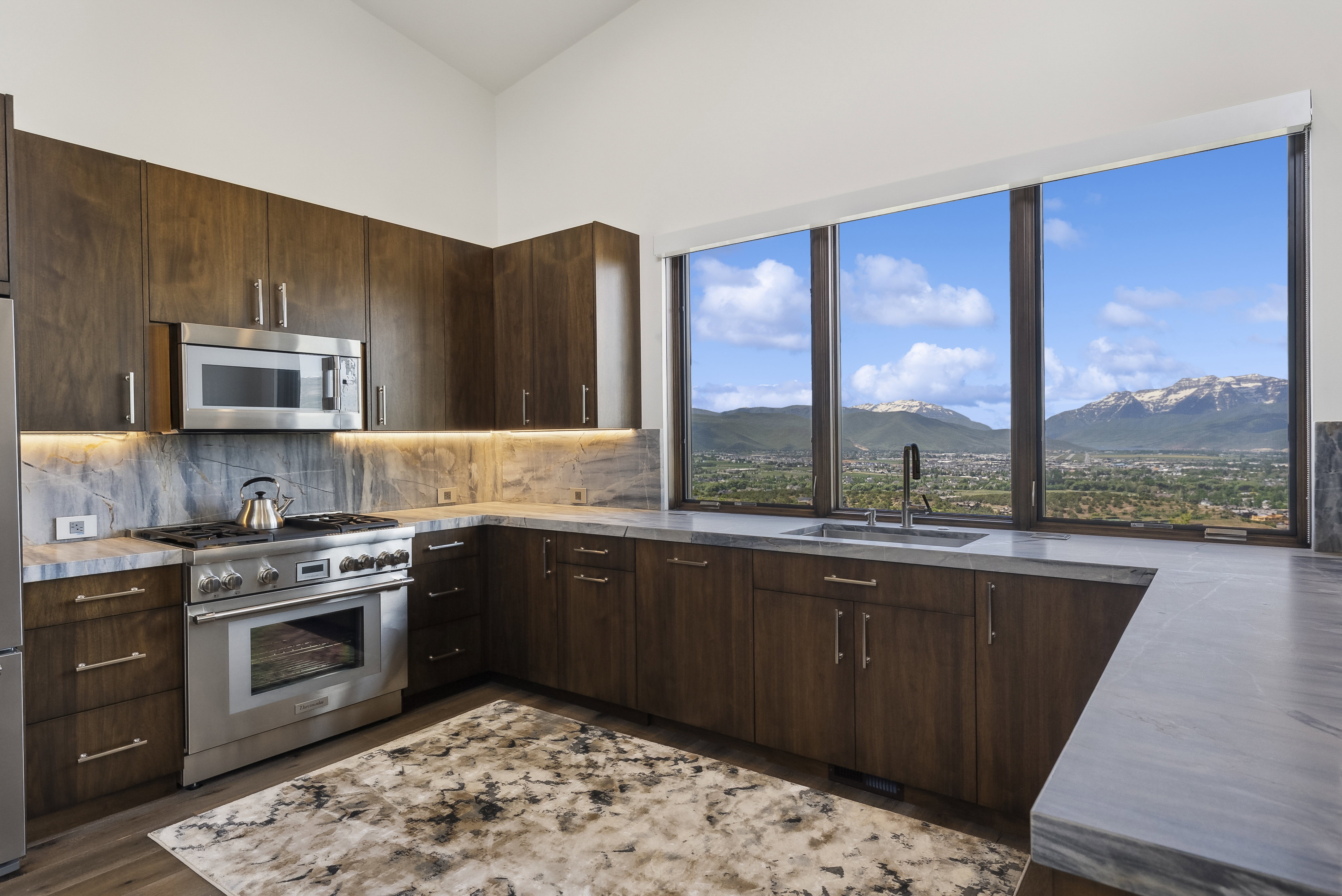 Kitchen featuring floor-to-ceiling windows with mountain scenery, rich wood cabinetry, stainless steel appliances, under-cabinet lighting, and stone countertops with matching backsplash