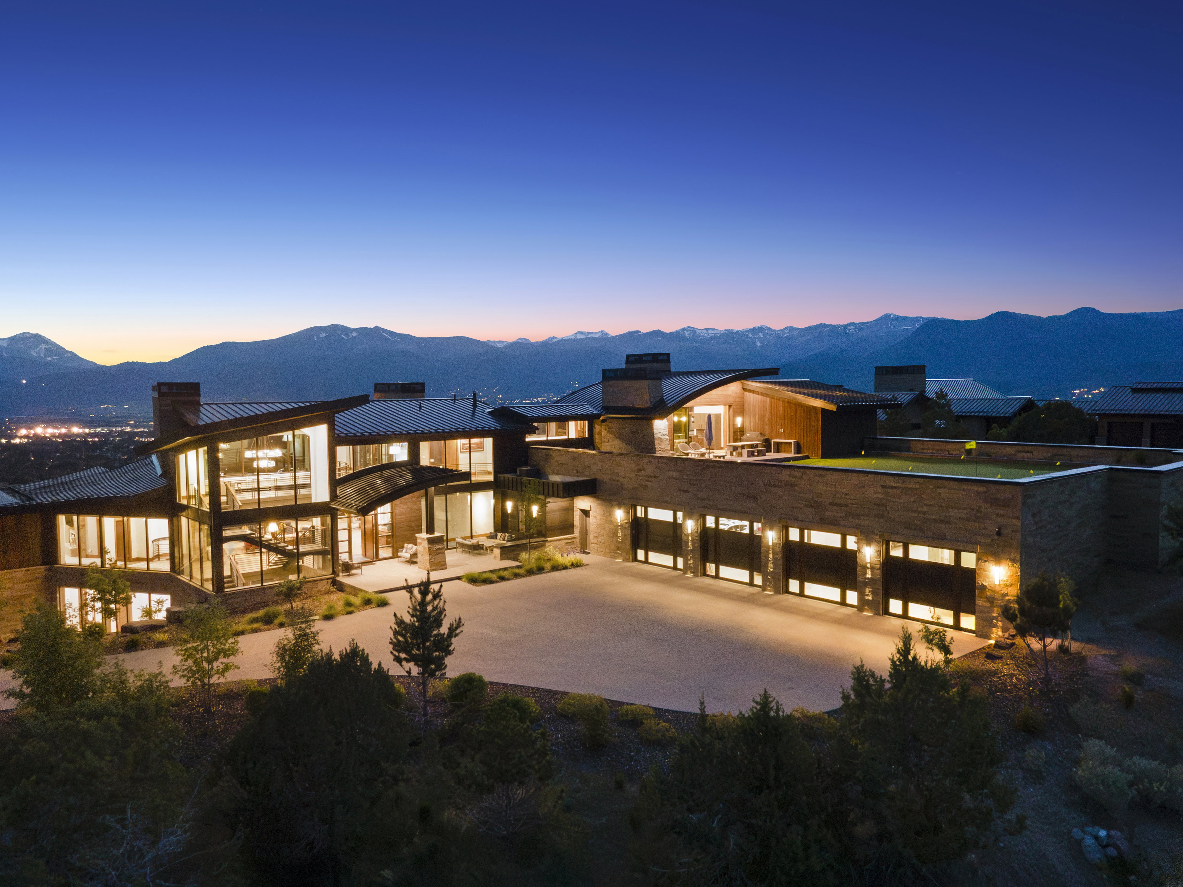 Contemporary residence featuring extensive floor-to-ceiling windows, a stone and wood siding exterior, multiple garage bays with frosted glass panels, an upper-level patio, and a mountain backdrop