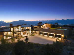 Contemporary residence featuring extensive floor-to-ceiling windows, a stone and wood siding exterior, multiple garage bays with frosted glass panels, an upper-level patio, and a mountain backdrop