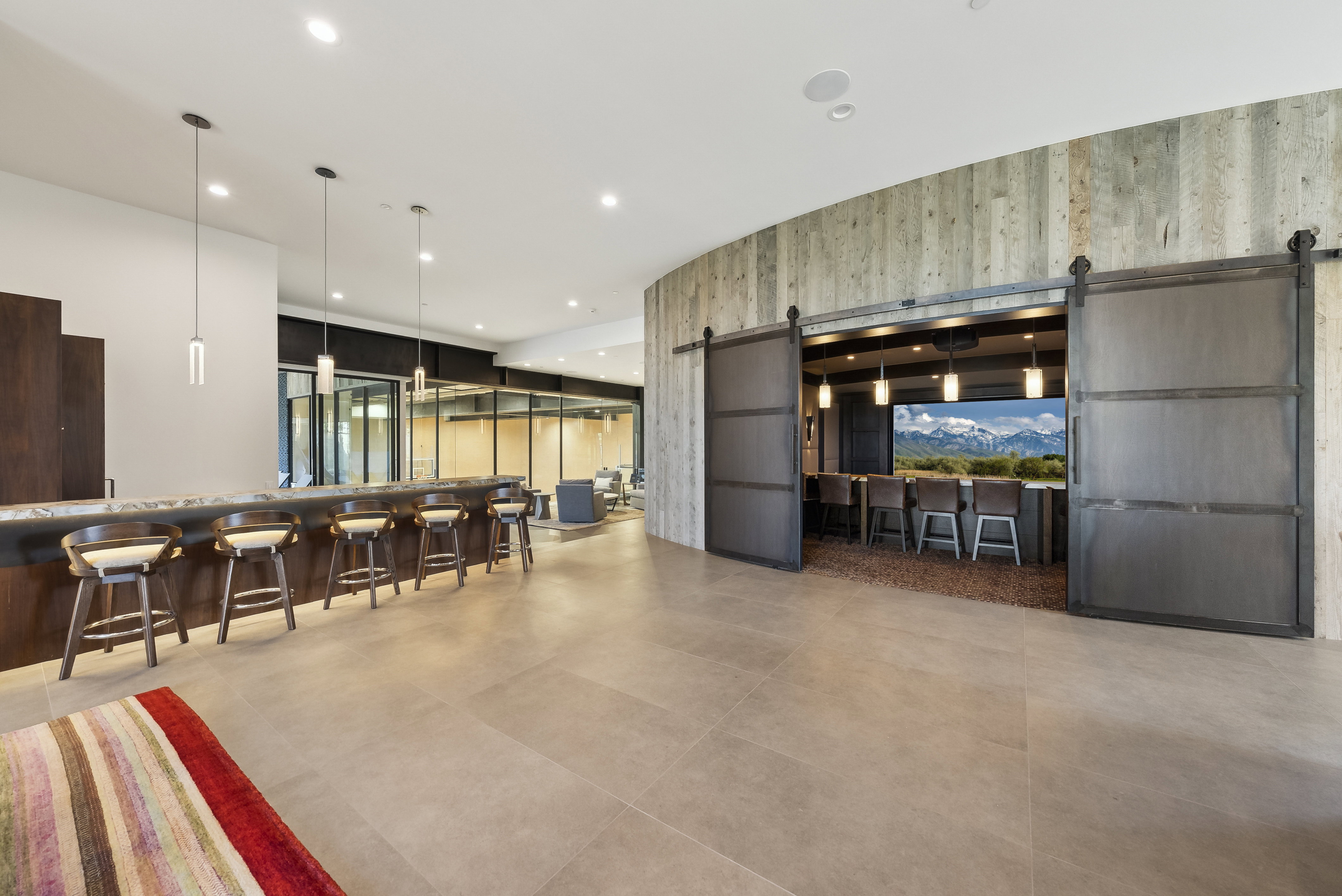 Spacious entertainment area featuring a long bar with pendant lighting, a curved wood-paneled accent wall, and a separate bar area behind sliding barn doors with a mountain landscape
