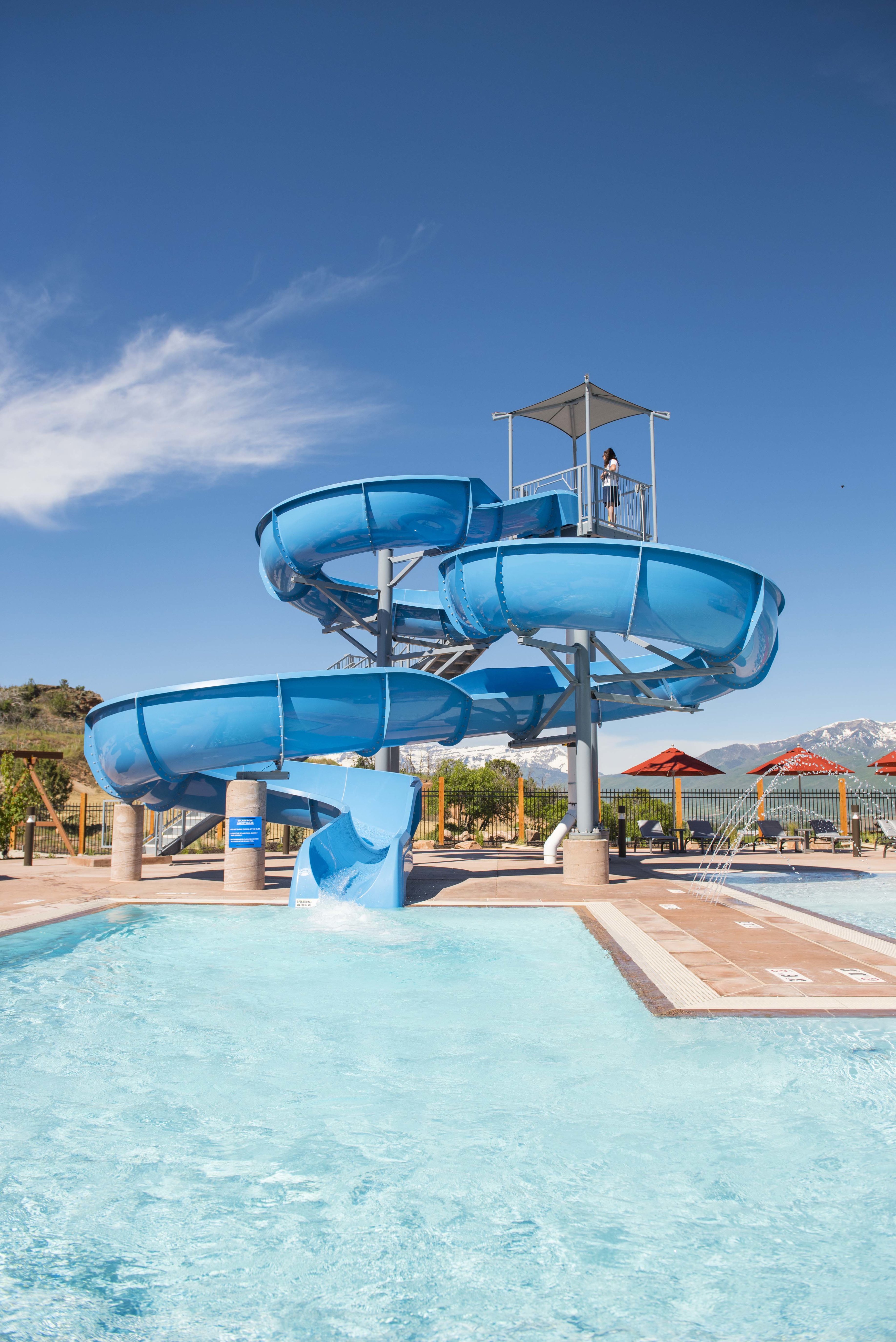 Resort-style aquatic amenity featuring a multi-story water slide structure with a shaded top platform, a pool with a tiled perimeter, and surrounding hardscaping with umbrellas
