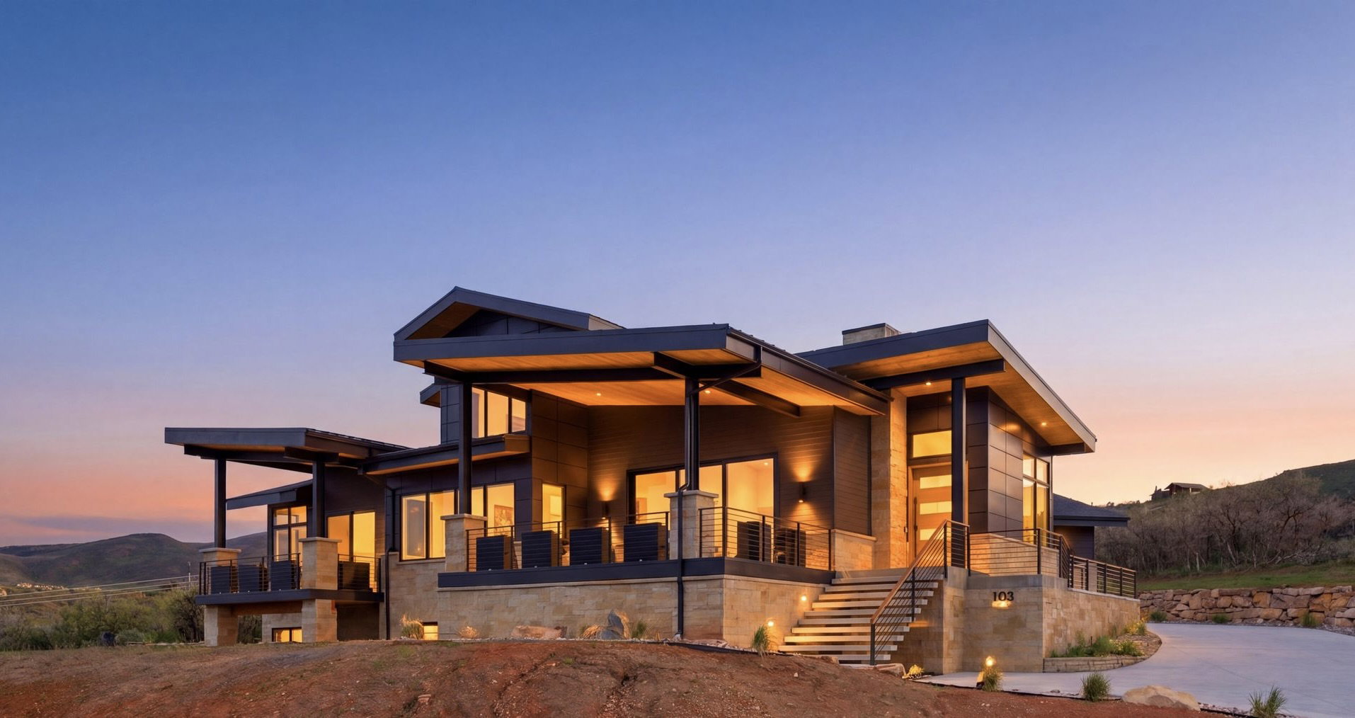 Modern architectural residence featuring multiple balconies, extensive exterior lighting, a stone-clad foundation, a multi-level entry staircase, and an asphalt driveway